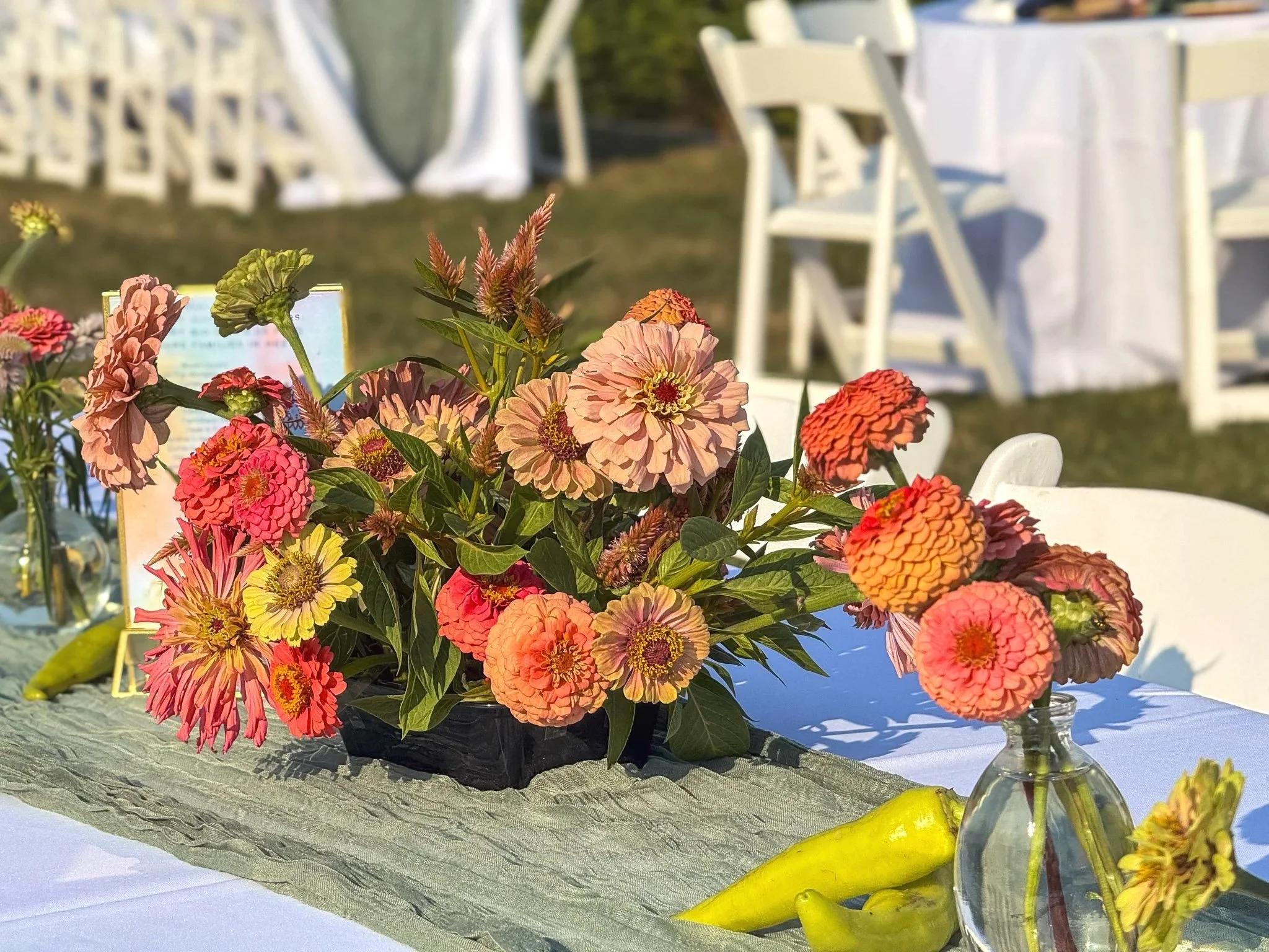 A floral centerpiece with pink, orange, and yellow flowers in a glass vase, set on a table with lemons and a lime. In the background, white chairs and tables are visible, suggesting an outdoor event.