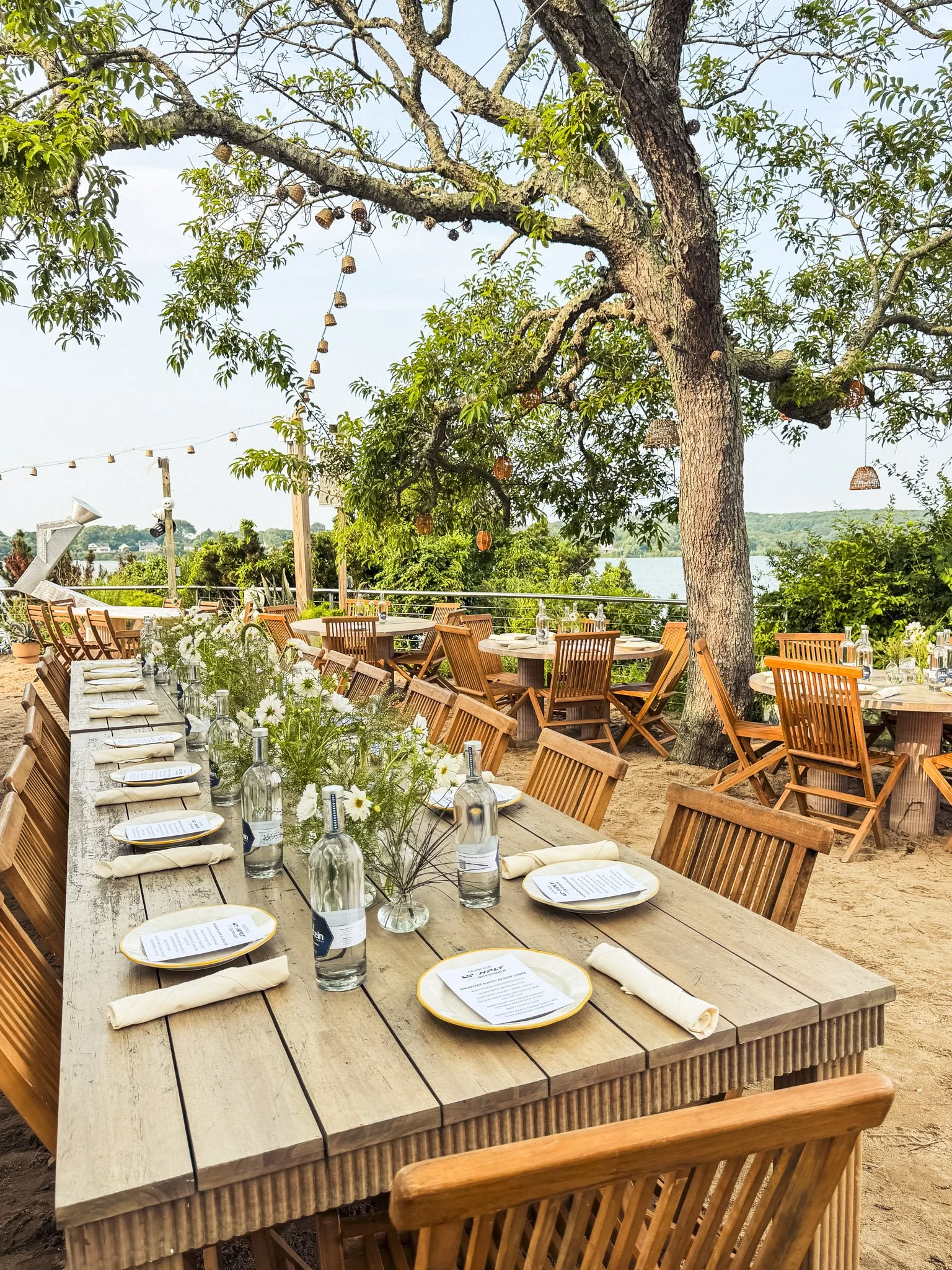 An outdoor dining setup with wooden tables and chairs under a large tree, with string lights and basket lanterns hanging. The tables are set with glasses, napkins, and menus, overlooking a body of water with lush greenery in the background.