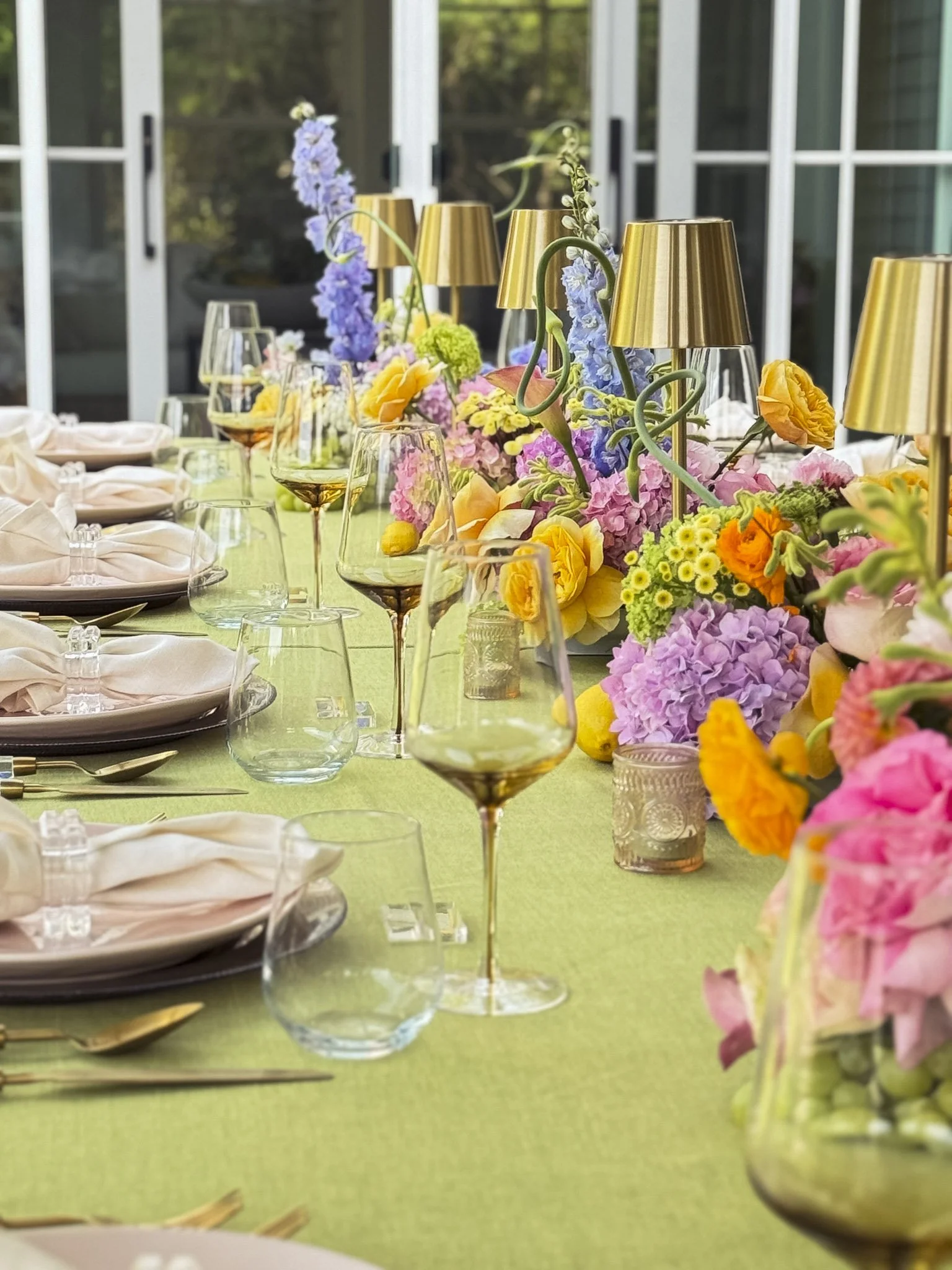 A table set with colorful flowers, wine glasses, and napkins, with a bright green tablecloth and a background of glass doors.