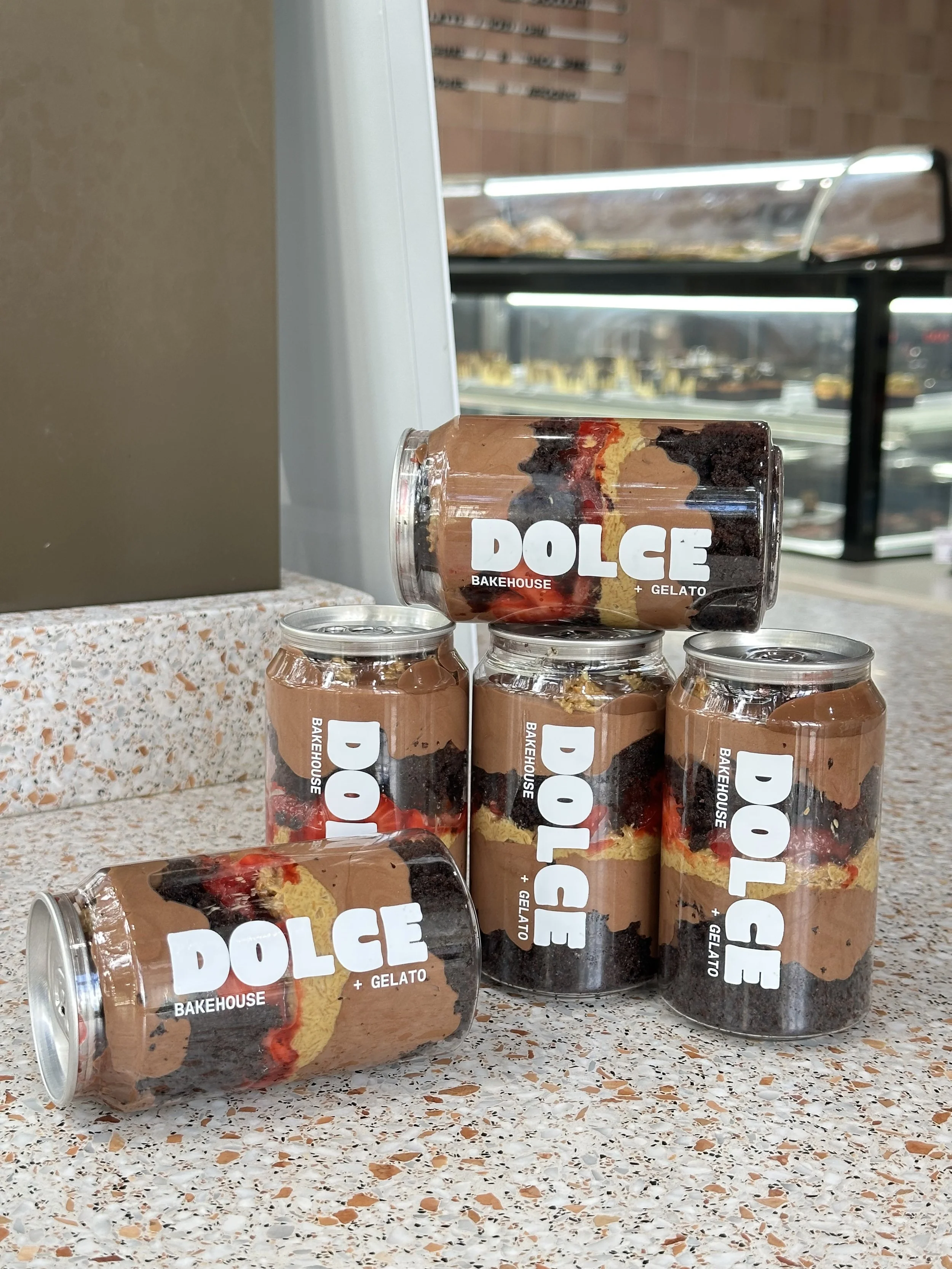 Multiple jars stacked on a speckled countertop display snack cakes labeled "DOLCE Bakehouse + Gelato". The backdrop includes a display of baked goods and a window showing a parking lot with cars.