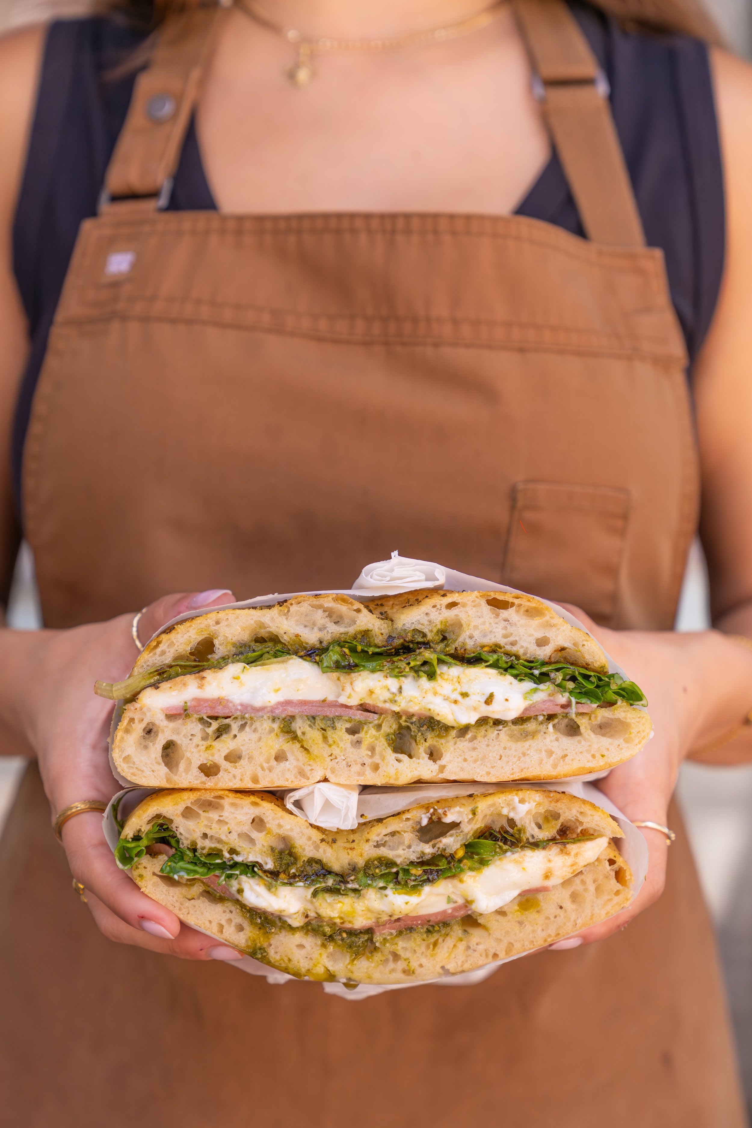 Staff is wearing an apron, holding one of the deli sandwiches.