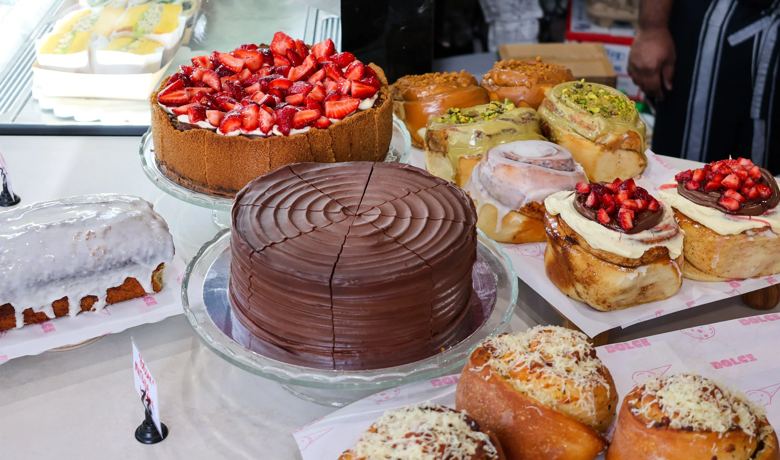 A variety of desserts including a strawberry-topped cake, a chocolate cake, and assorted pastries with toppings like pistachios and whipped cream on display at a bakery or cafe.