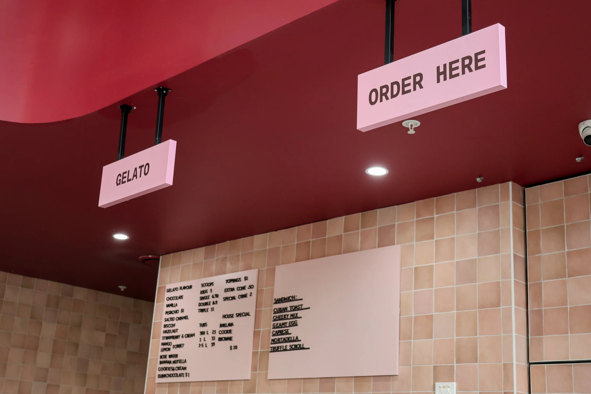 Pink hanging signs that read 'Gelato' and 'Order Here' in a dessert shop with tiled walls.