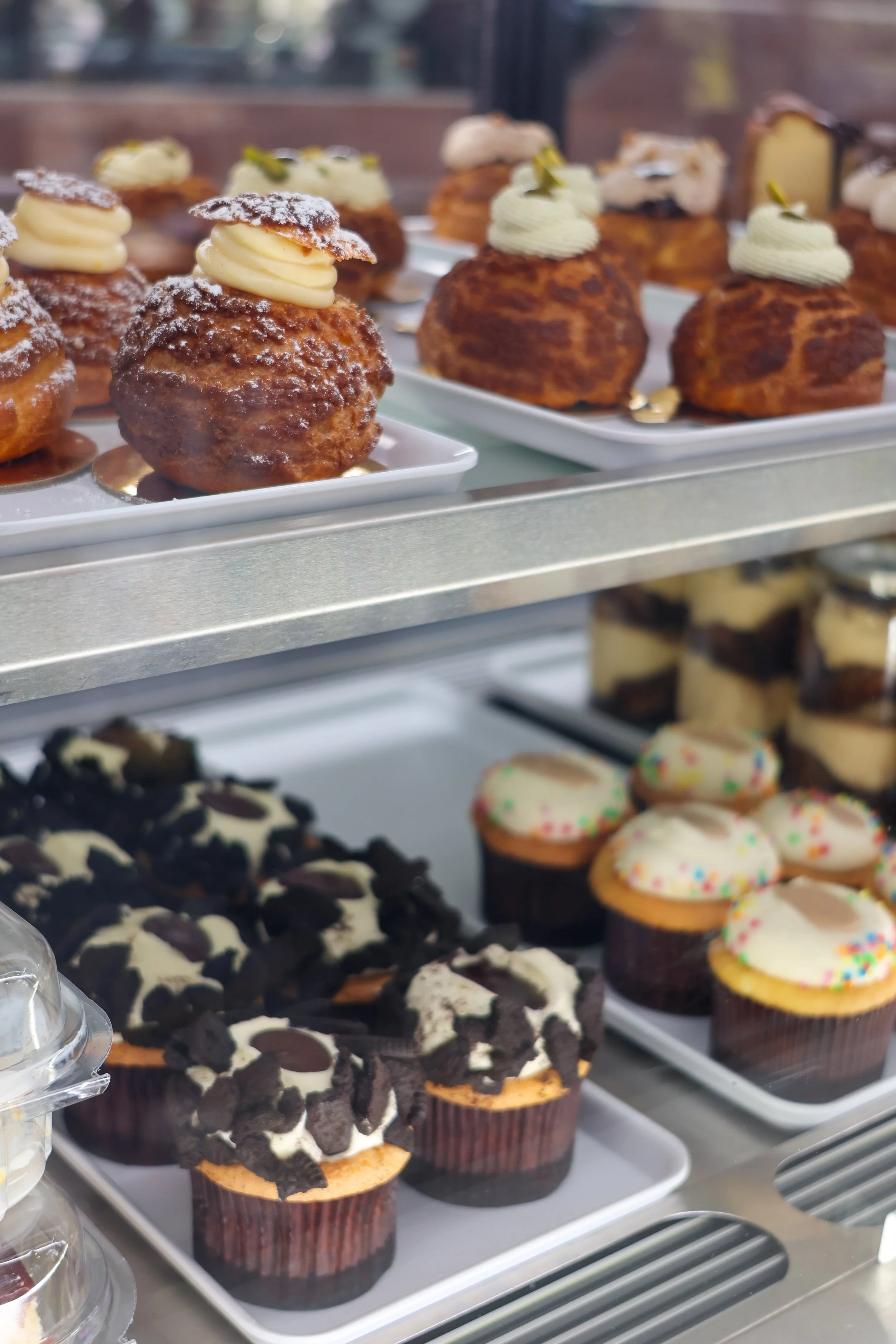 Assorted cupcakes with various toppings displayed on trays in a bakery. Some have frosting and sprinkles, others have cookies or other decorations.