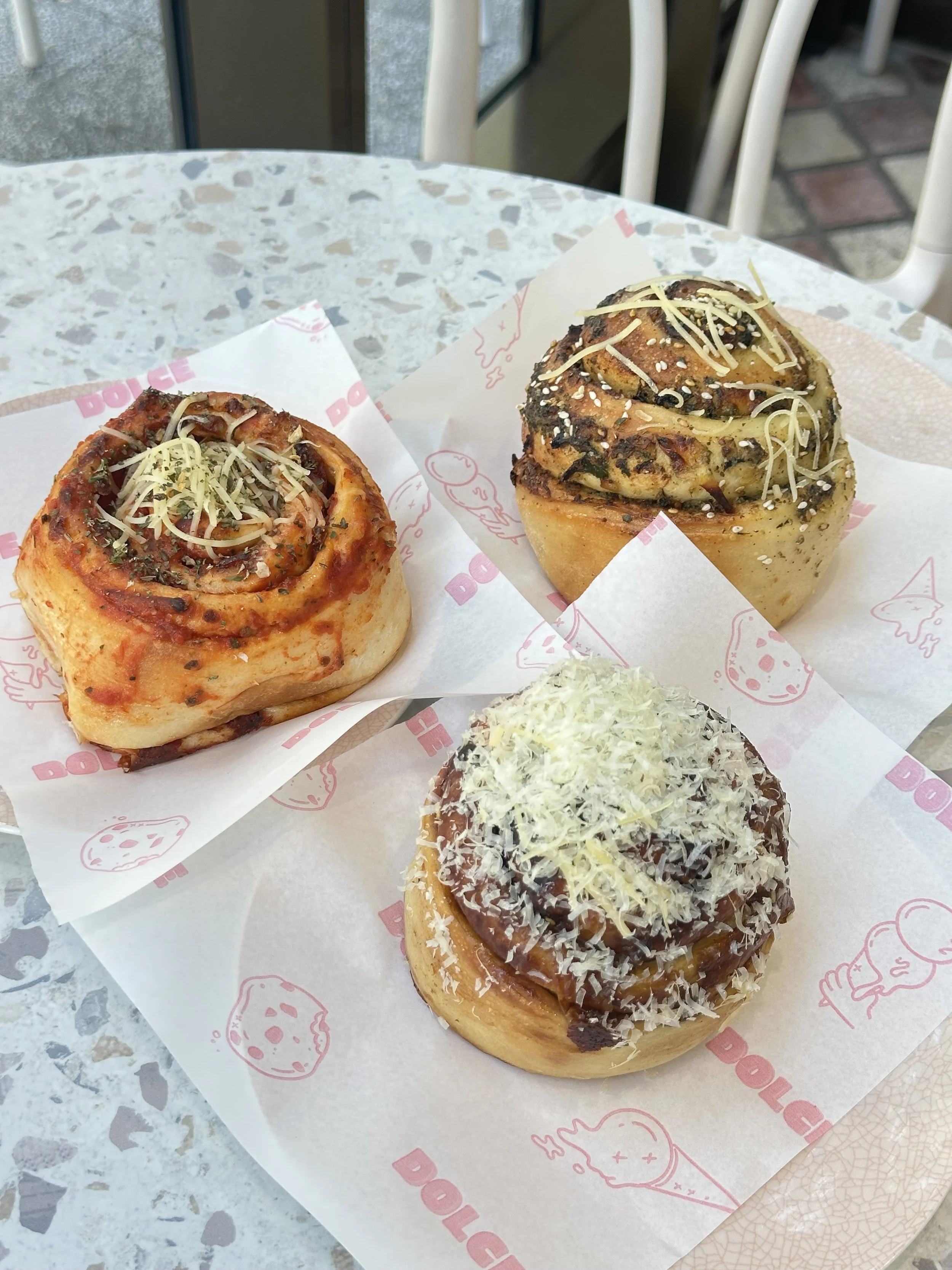 Three cinnamon rolls topped with icing, shredded cheese, and sprinkles on branded paper placed on a table outside.