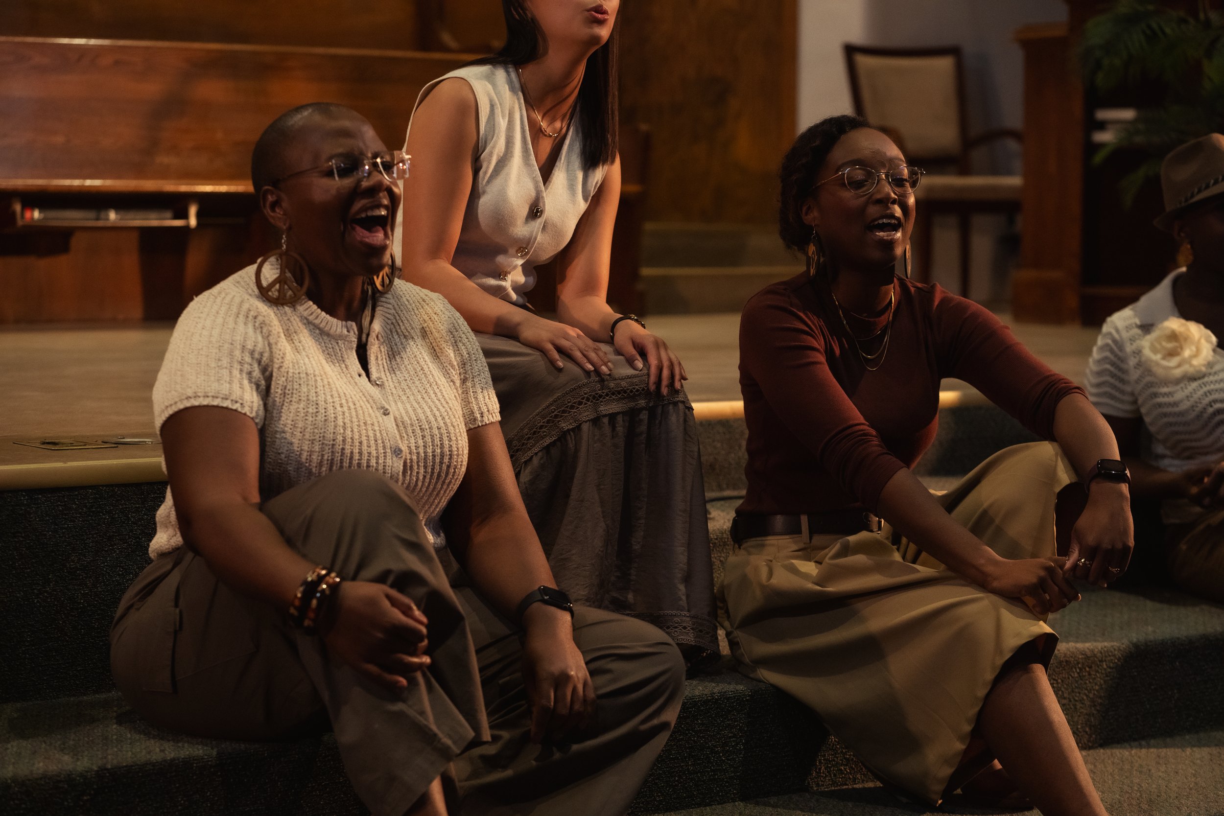 Four women sitting and standing in a dimly lit room, laughing and singing together, with wooden décor and chairs in the background.