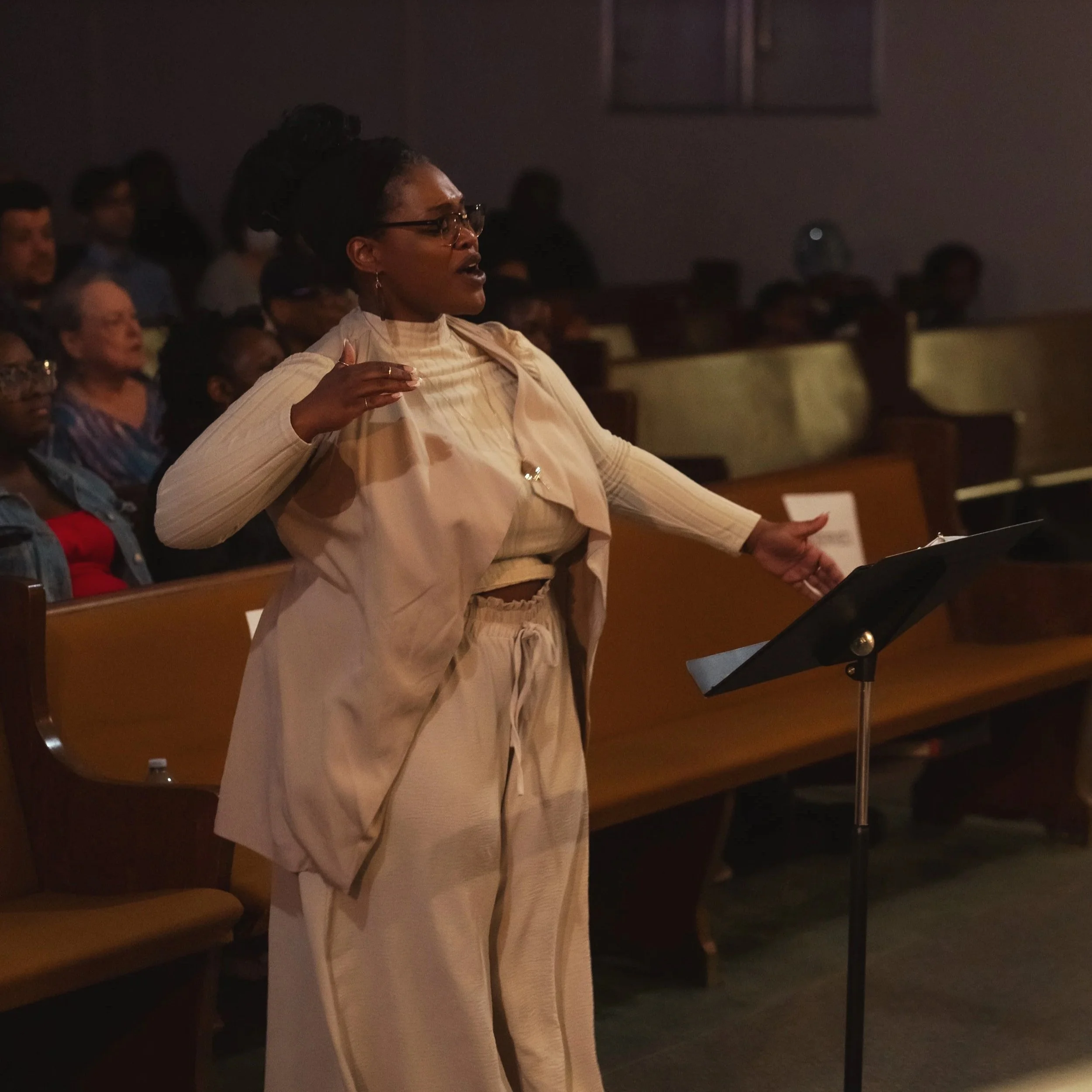 A woman conducting in a dark, indoor setting, possibly a concert or choir rehearsal, with several people seated in the background.