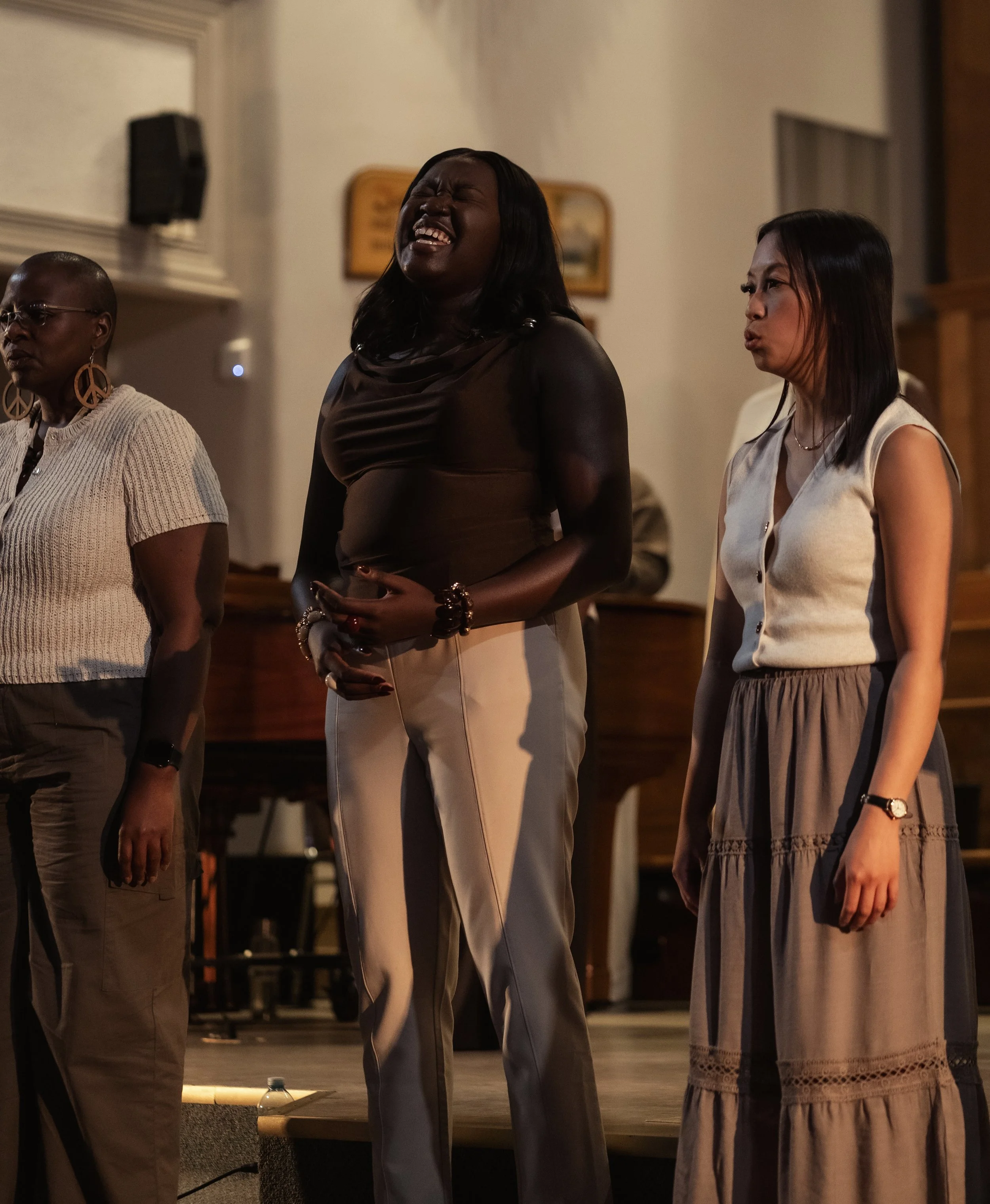 Three women standing in a room, two of them appear to be singing or speaking, one is wearing earrings with peace signs, and another is wearing a white sleeveless top with lace detail.