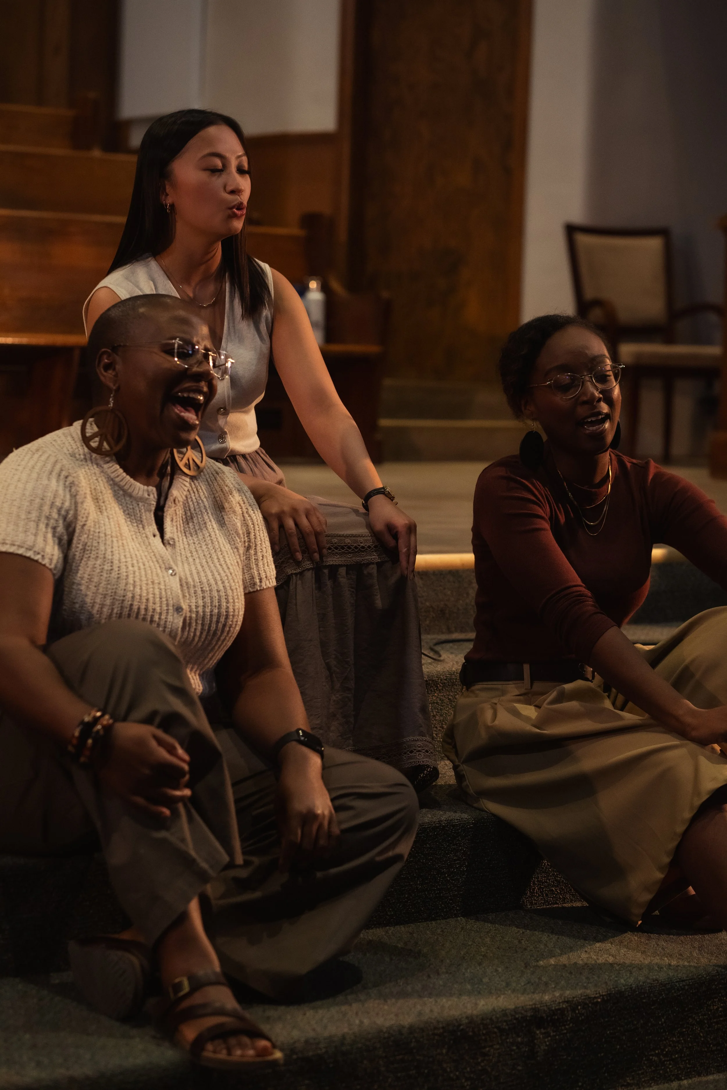 Three women sitting on the floor in a room, singing or praying with closed eyes, and one standing woman singing with eyes closed, in a dimly lit setting.