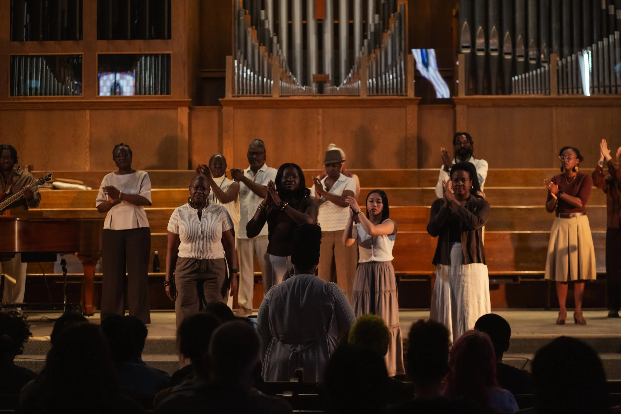A diverse group of people performing on stage in a concert hall, with some clapping and others standing, while an organ is visible in the background and an audience watches.