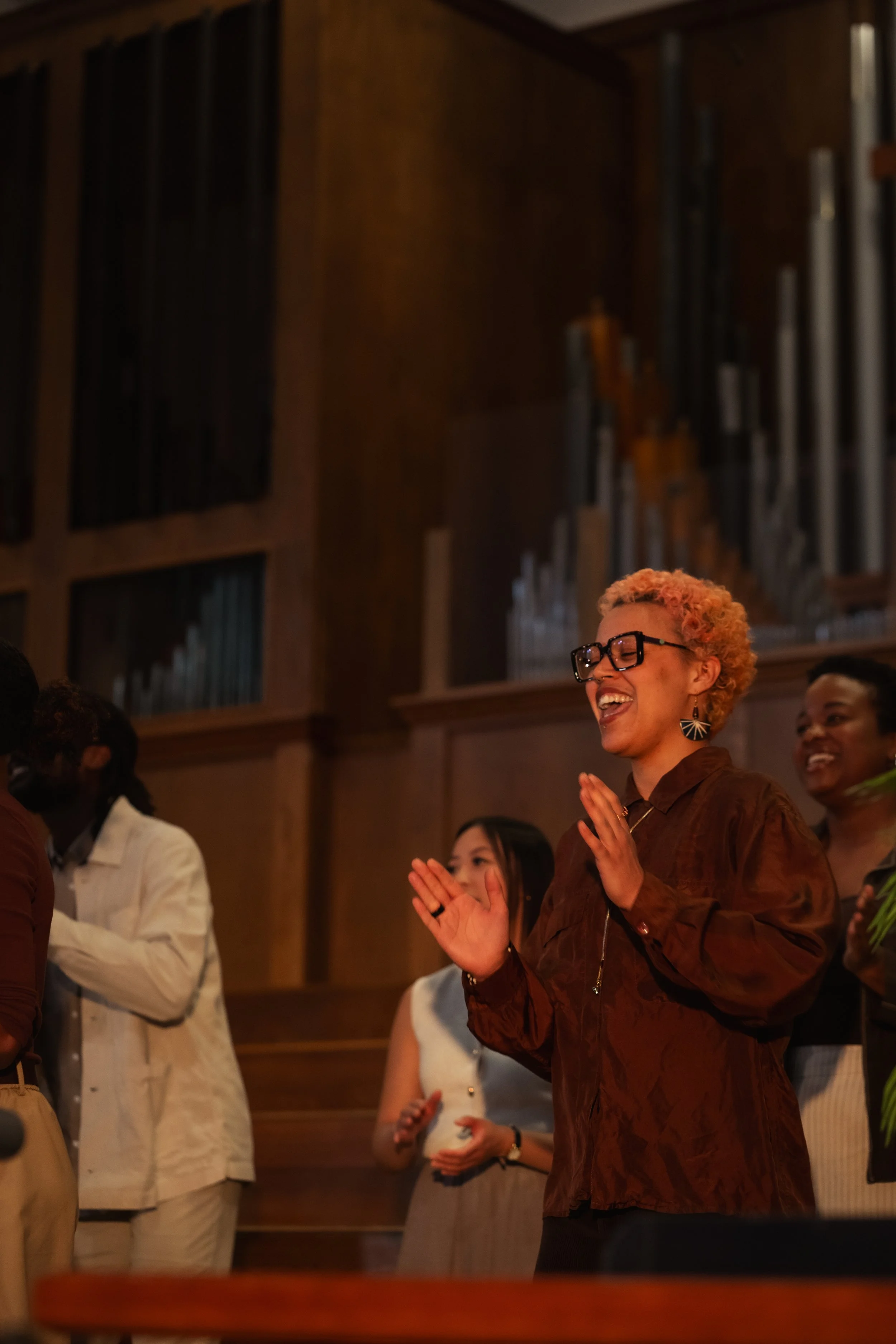 A group of people smiling and clapping in a warmly lit room with wooden panel walls and a large pipe organ in the background.