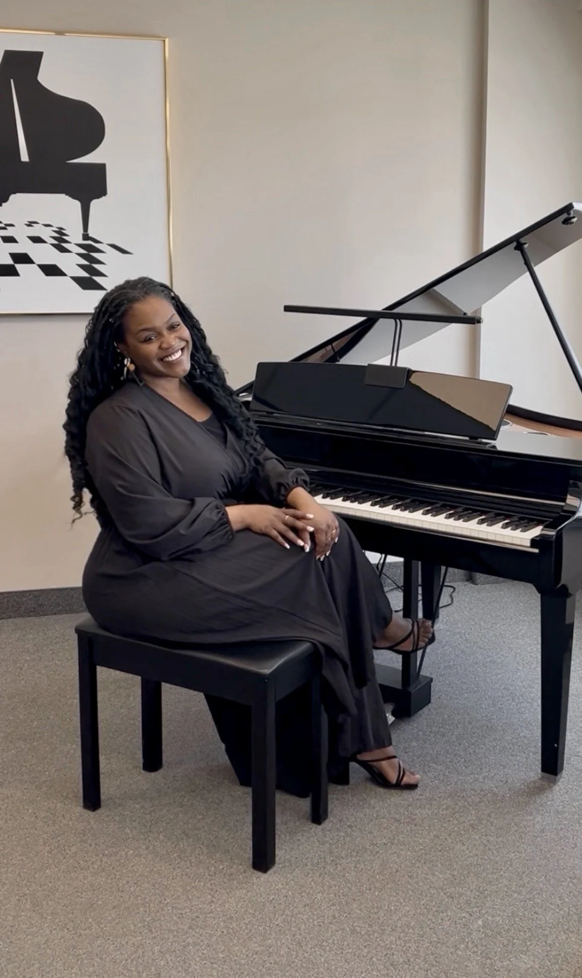 A woman sitting next to a grand piano, smiling, in a room with beige walls and wall art of a black piano silhouette.