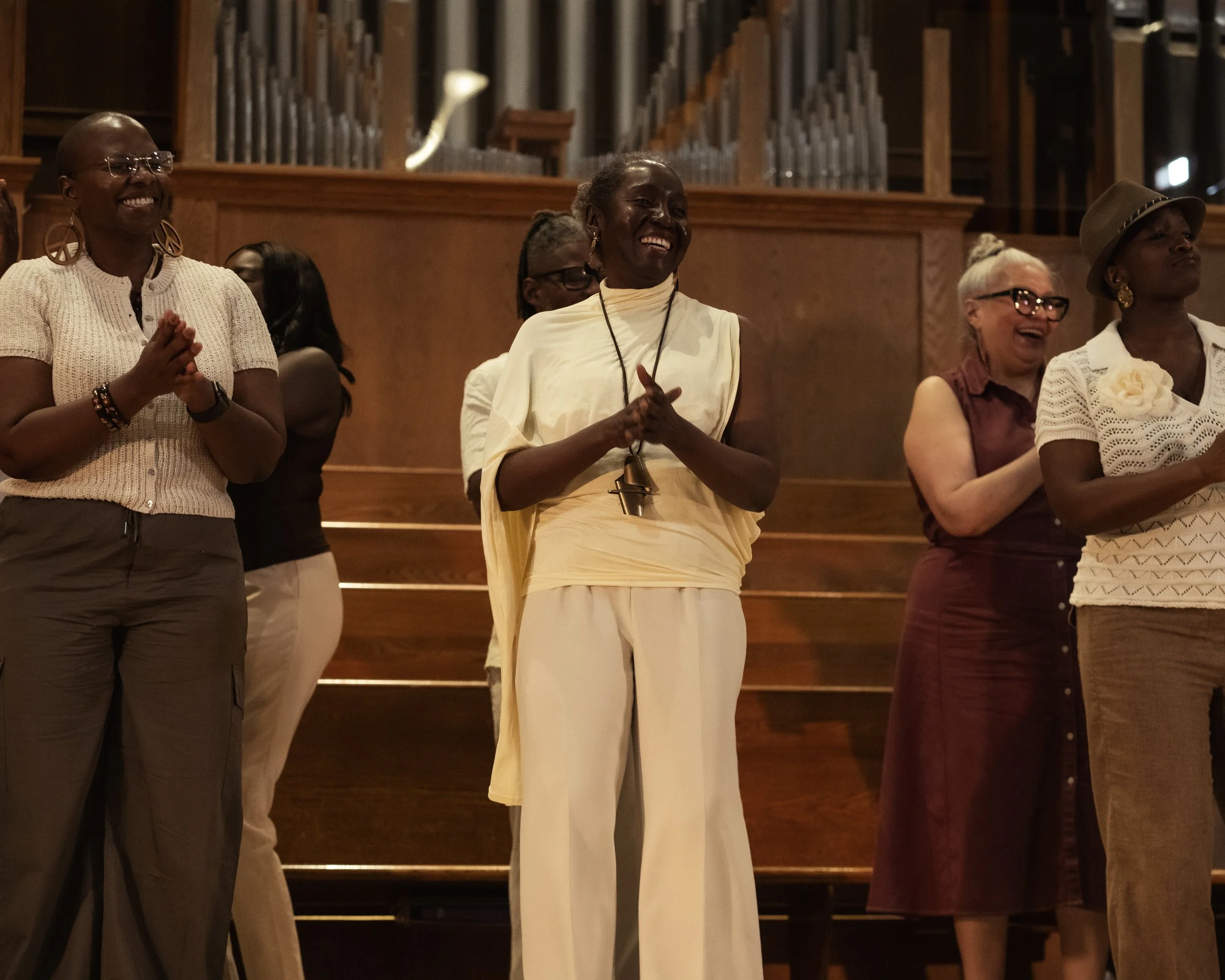 A diverse group of women standing on a stage, smiling and clapping, in a church or concert hall with wooden architecture and pipe organ pipes in the background.