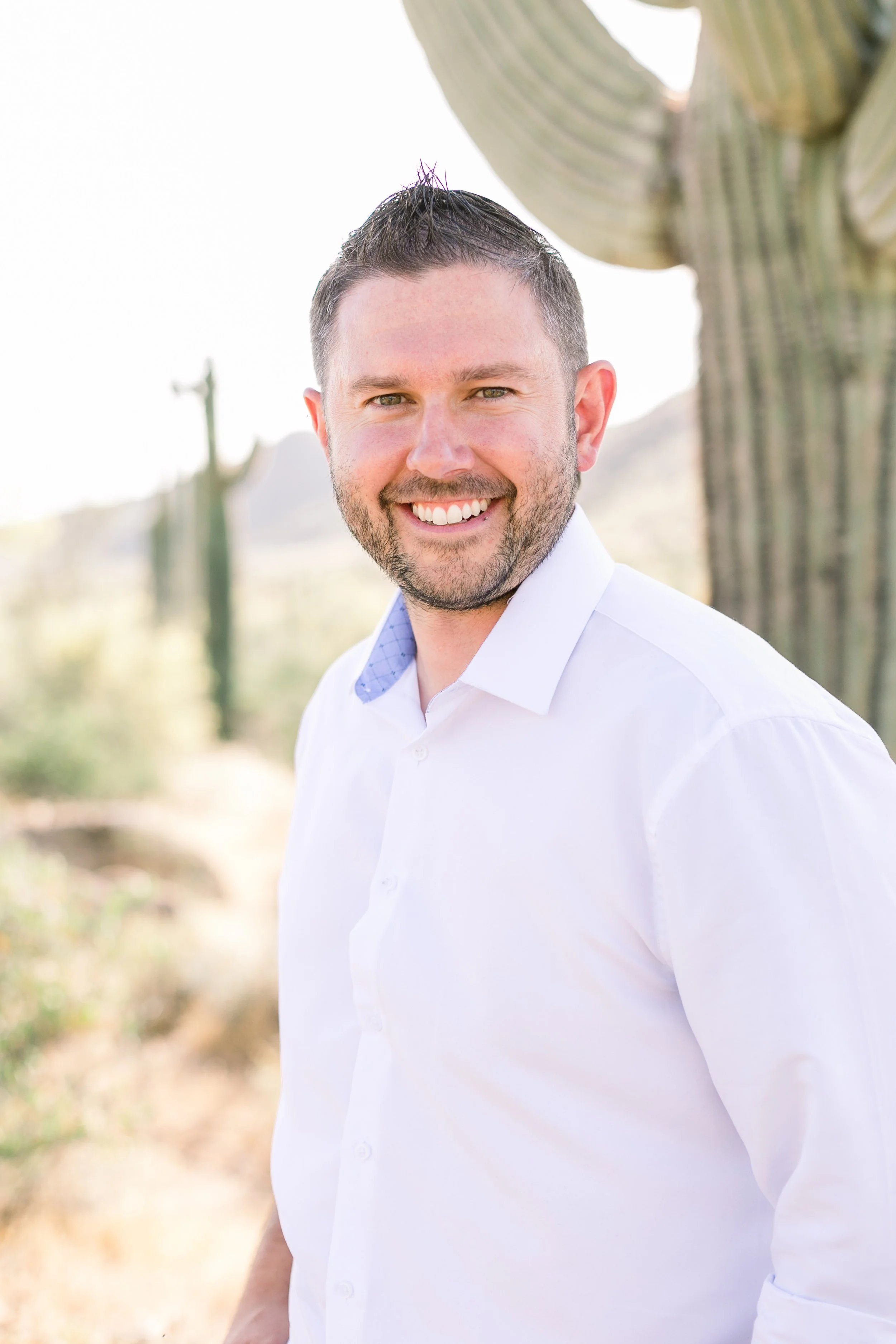 A smiling man in a white shirt standing outdoors with desert plants and cacti in the background.