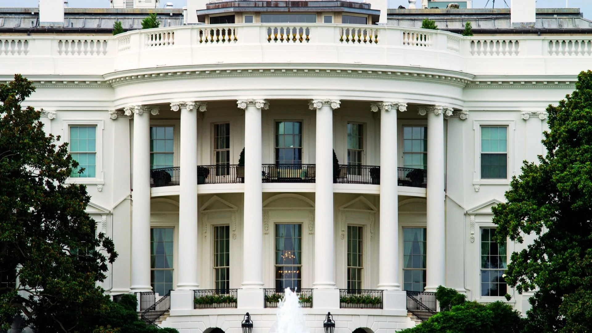 The White House facade with tall white columns, multiple windows, trees on both sides, and a small fountain in front.