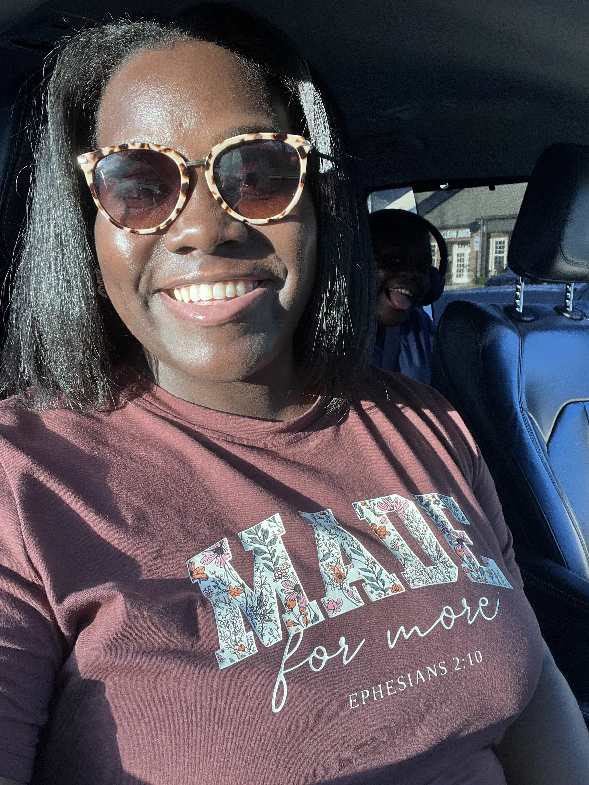 A woman with dark hair and glasses smiling inside a vehicle, wearing a burgundy T-shirt with floral lettering that says "AMAZE" and a Bible verse reference, Ephesians 2:10.