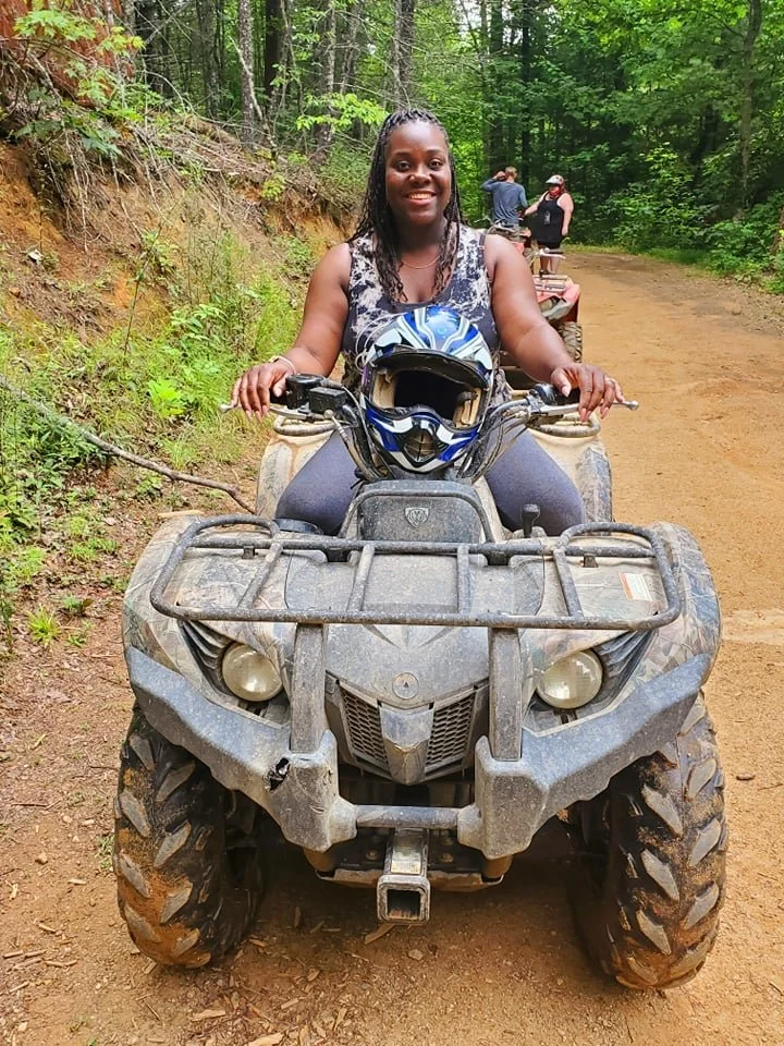 A woman is sitting on a dirt bike in a wooded outdoor trail area. She is smiling and wearing a sleeveless top, with a blue and white helmet resting on the bike's handlebar. In the background, other people are seen near parked ATVs.