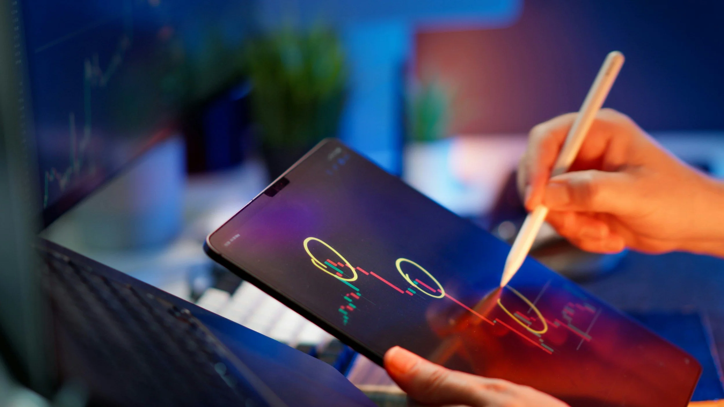 Persona usando un stylus en una tableta con gráficos y datos financieros en la pantalla, en un entorno de oficina con una computadora portátil y pantalla grande en el fondo.