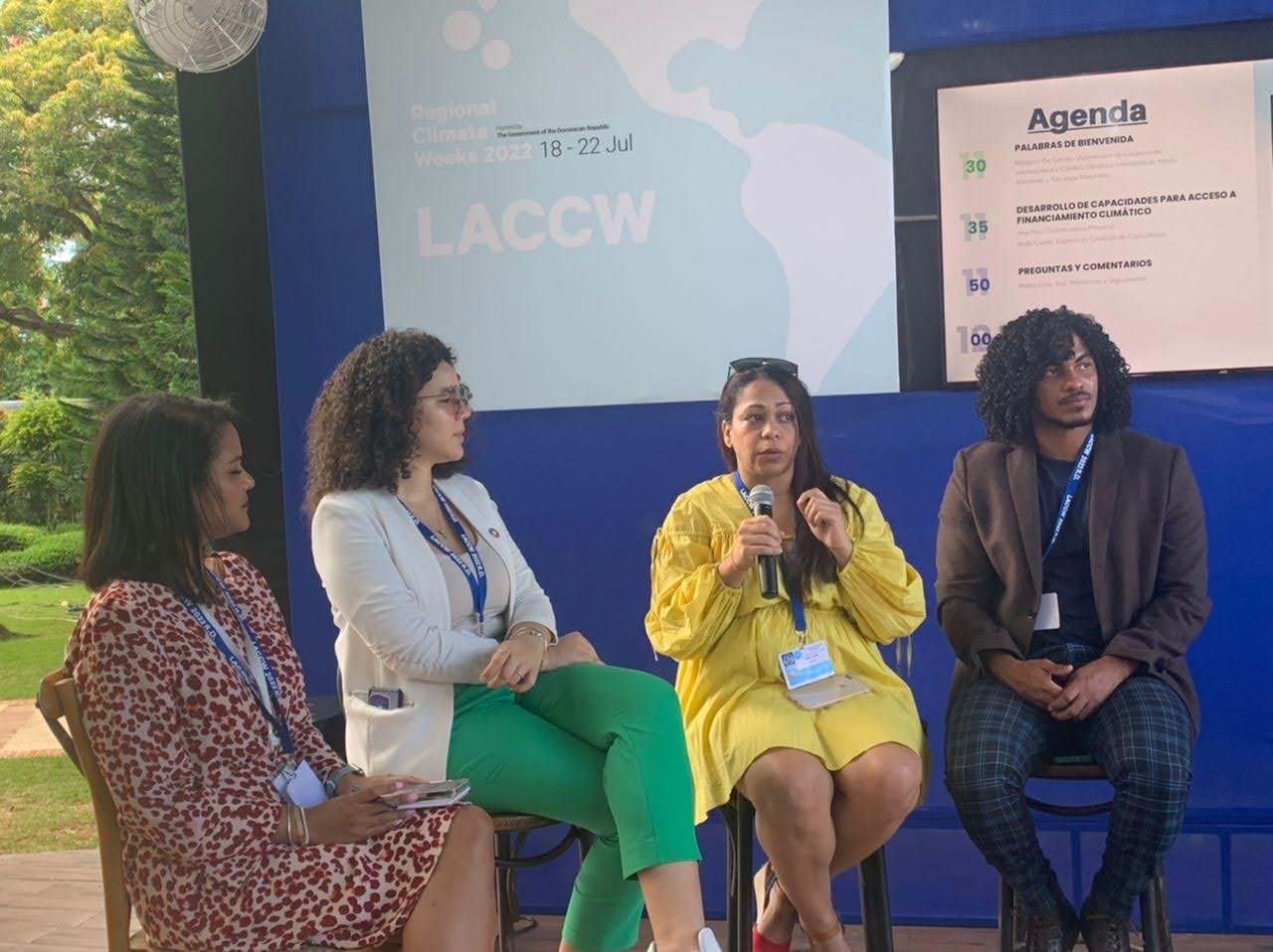 Tres mujeres y un Joven participation en un panel de discusión durante el evento 'Semana Regional del Clima 2022', en un escenario con fondo azul y una pantalla con programa de agenda visible.