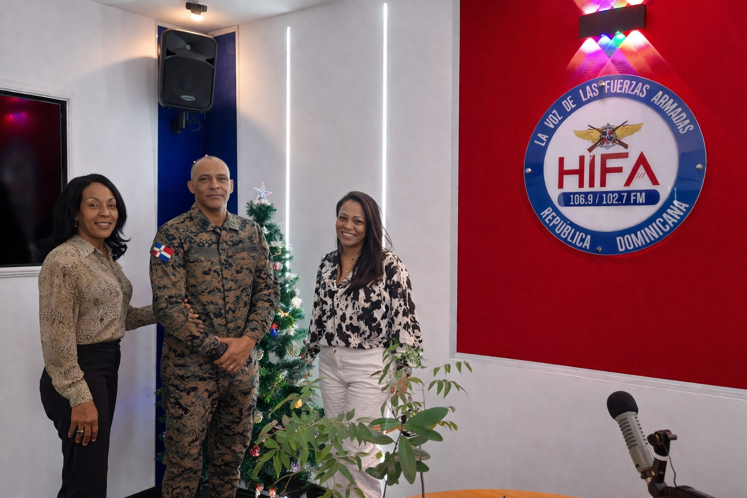 Tres personas posando en una cabina de radio con un árbol de Navidad decorado y un logotipo de Radio HIFA en la pared, en la República Dominicana.