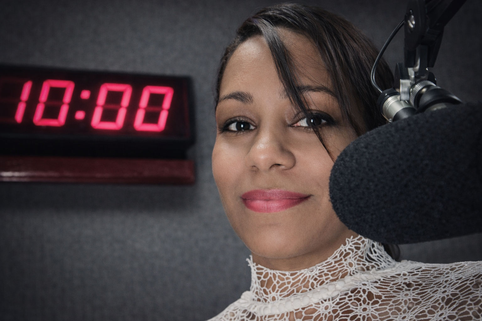 Una mujer en un estudio de radio frente a un micrófono, con un reloj digital que indica las 10:00, sonriendo y mirando hacia la cámara.