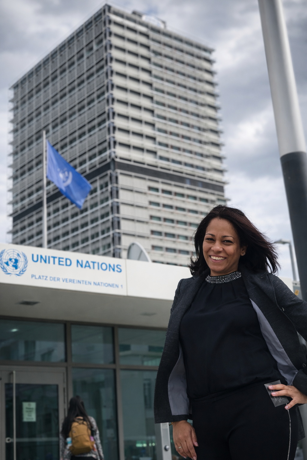 Mujer sonriendo frente a la entrada de las Naciones Unidas en Bonn, Alemania con un edificio alto y una bandera en el fondo.