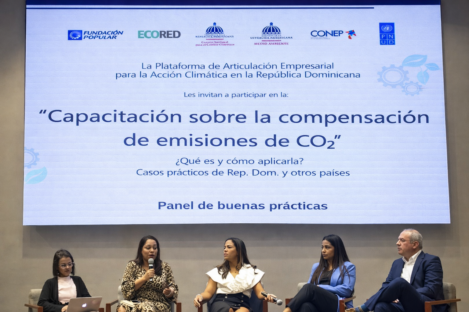 Un panel de cinco personas participando en una conferencia sobre compensación de emisiones de CO₂ en un auditorio, con una pantalla grande detrás que muestra información en español sobre la capacitación y buenas prácticas en República Dominicana.