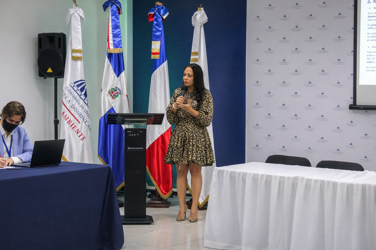 Mujer dando una conferencia frente a varias banderas, entre ellas la bandera de la República Dominicana, en un salón de reuniones con una pantalla proyectando texto.