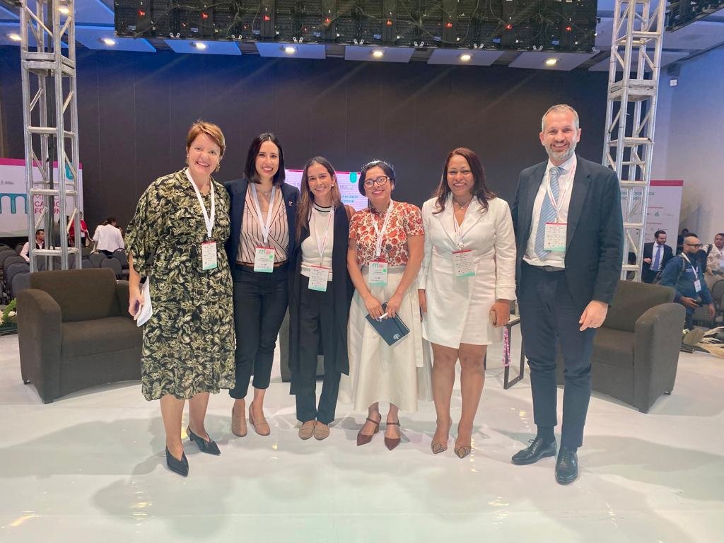 Grupo de siete personas en un escenario en un evento o conferencia, son mujeres y un hombre, vestimenta formal, fondo con pantallas y sillas.