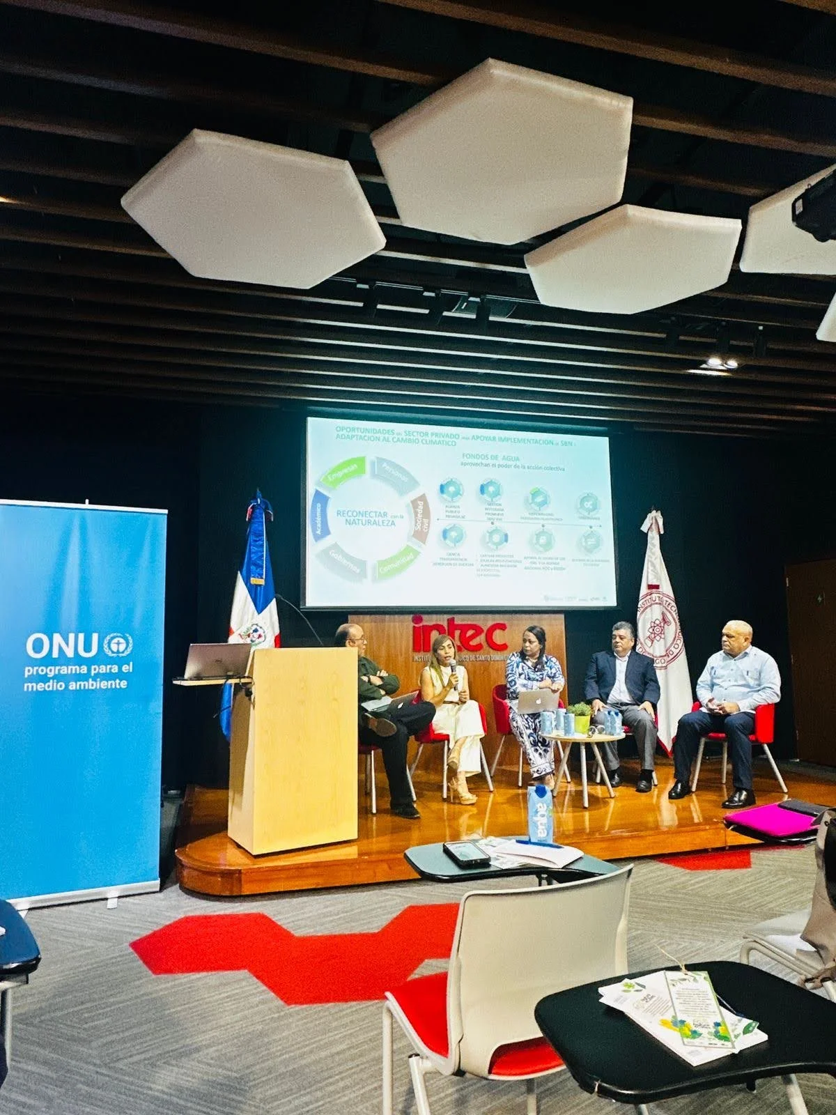 Panel de discusión en una conferencia sobre medio ambiente, con cinco personas sentadas en una pequeña mesa frente a un escenario, donde hay un proyector y un cartel de ONU