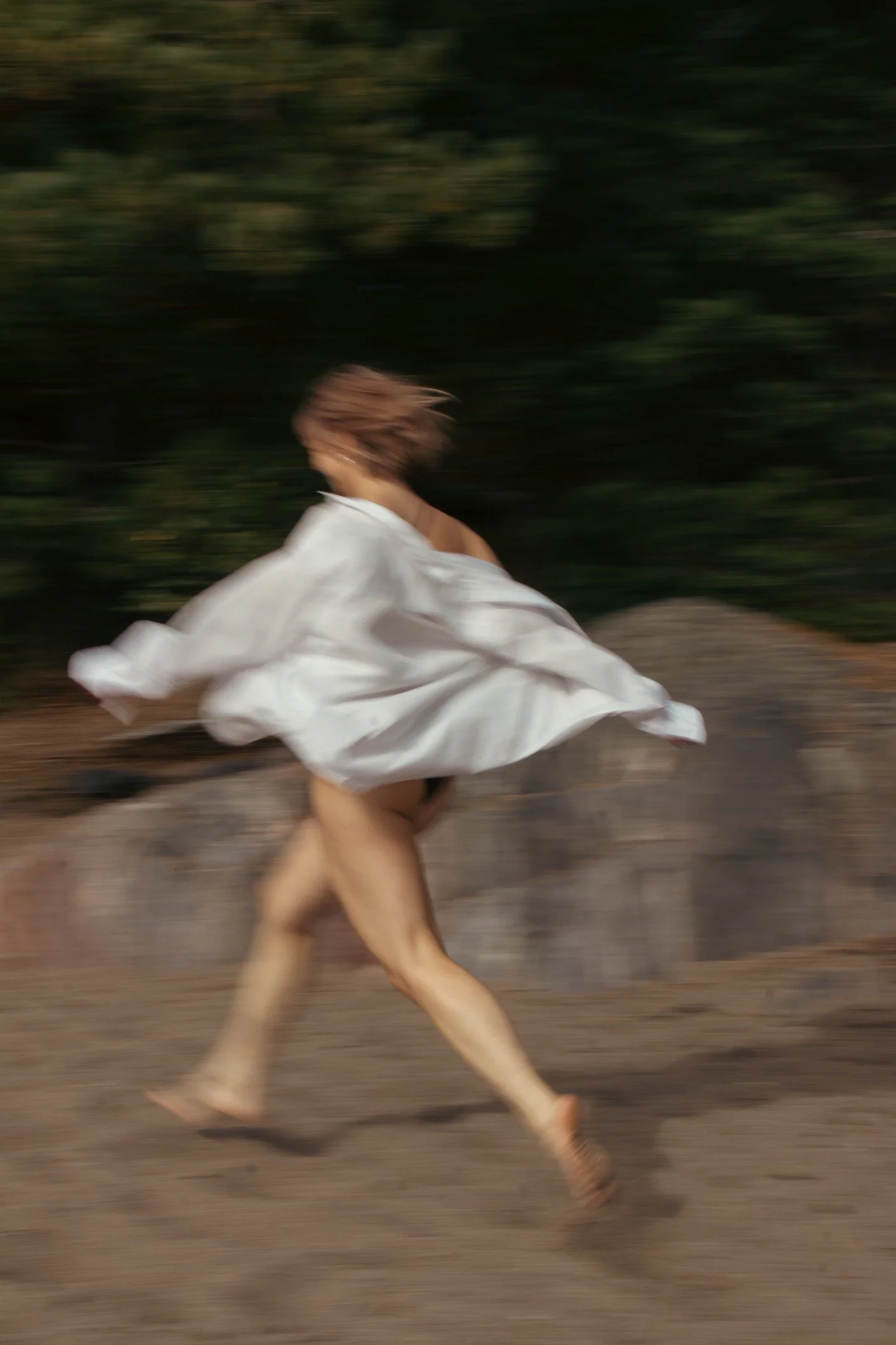 A person running outdoors at dusk wearing a white flowing dress, with a blurred background of trees and dirt ground.