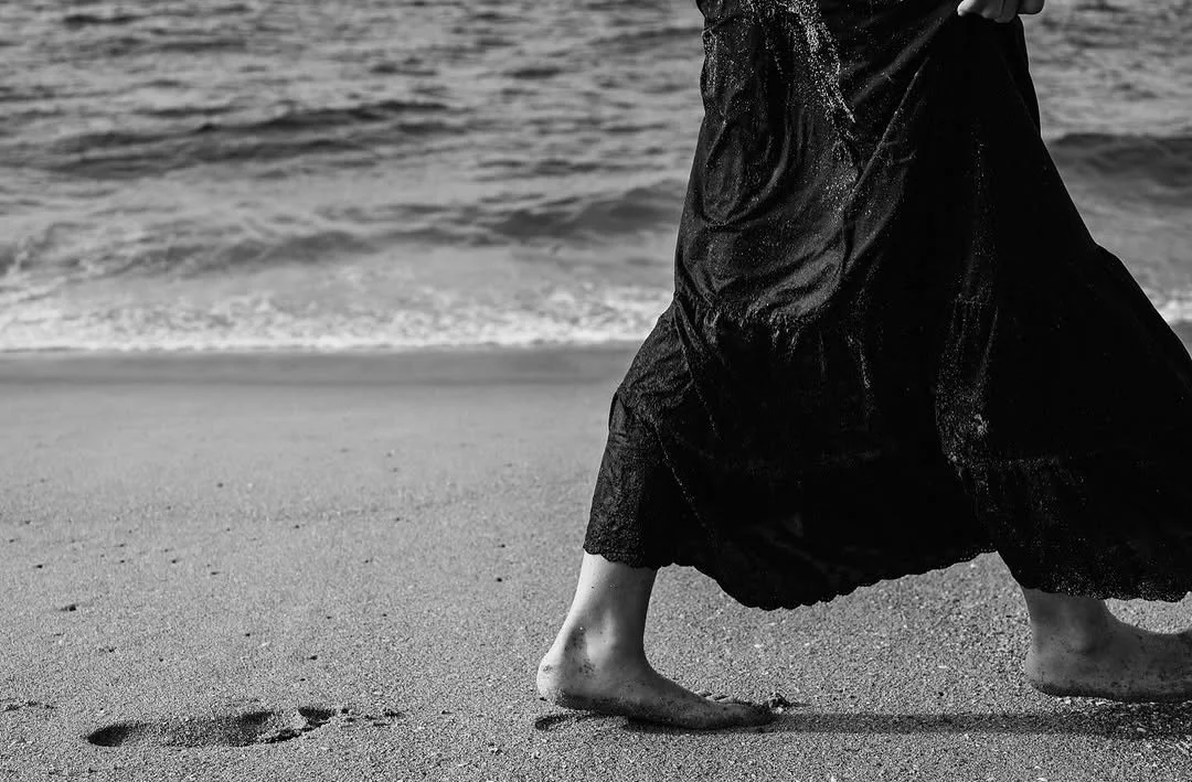 A person barefoot walking on the beach with black wide-legged pants, near the shoreline with waves in the background.