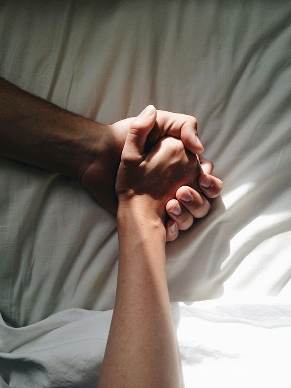 A person with light skin is holding hands with another person with darker skin, both resting their hands on a bed with white sheets.