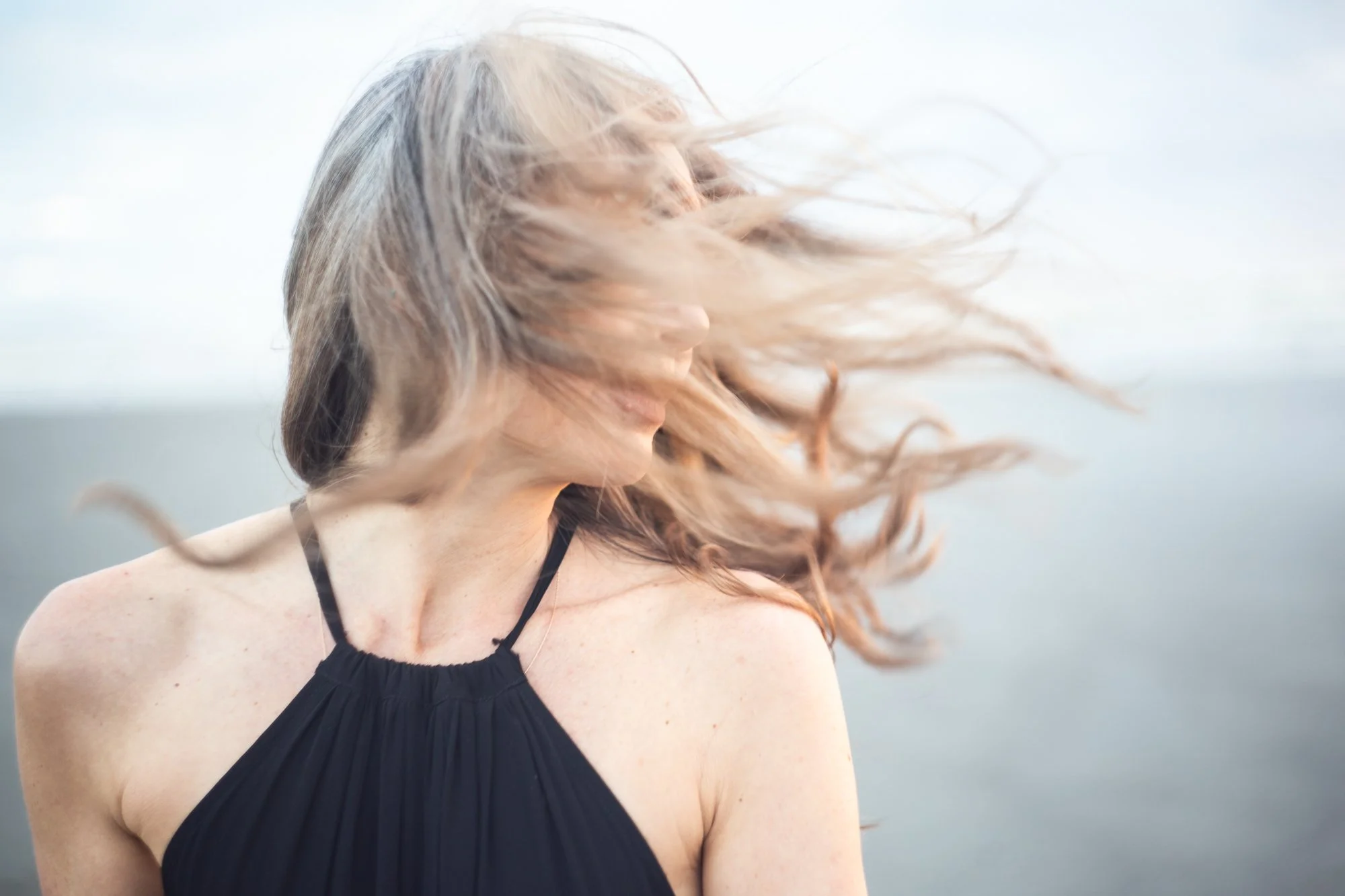 A woman with long reddish hair blowing in the wind is photographed outdoors against a soft, pale sky background. She is wearing a black, sleeveless top with thin straps.
