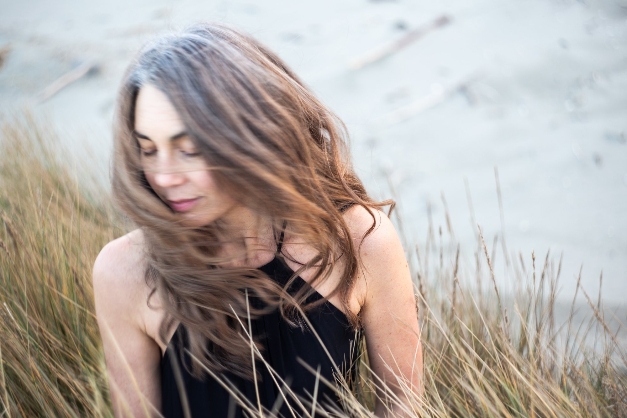 A woman with long brown hair sitting in a grassy field near water, looking down with a peaceful expression.