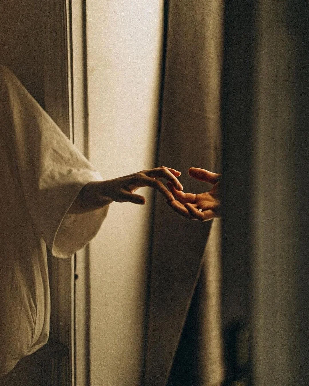 Two hands nearly touching, emerging from a bed and a curtain in a dimly lit room.