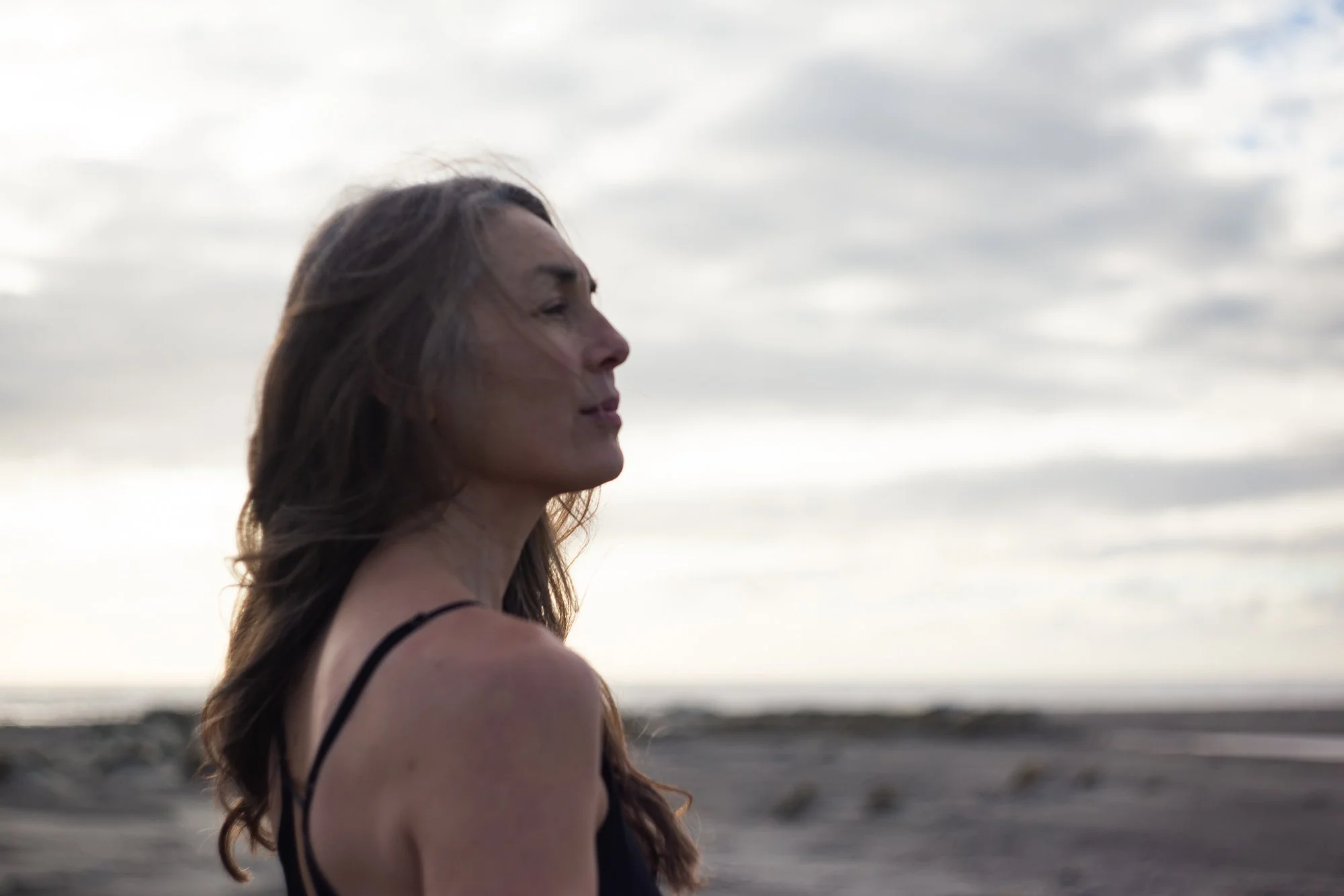 Side profile of a woman with long brown hair, wearing a black strap dress, standing on a beach with waves in the background, under cloudy sky.