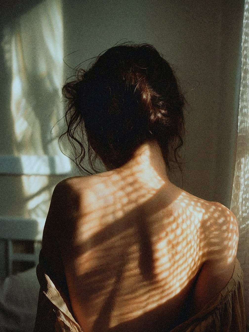 A woman with dark, curly hair seen from behind, sitting with her back exposed. Sunlight passing through a textured window creates a pattern of light and shadow on her skin and the wall behind her.