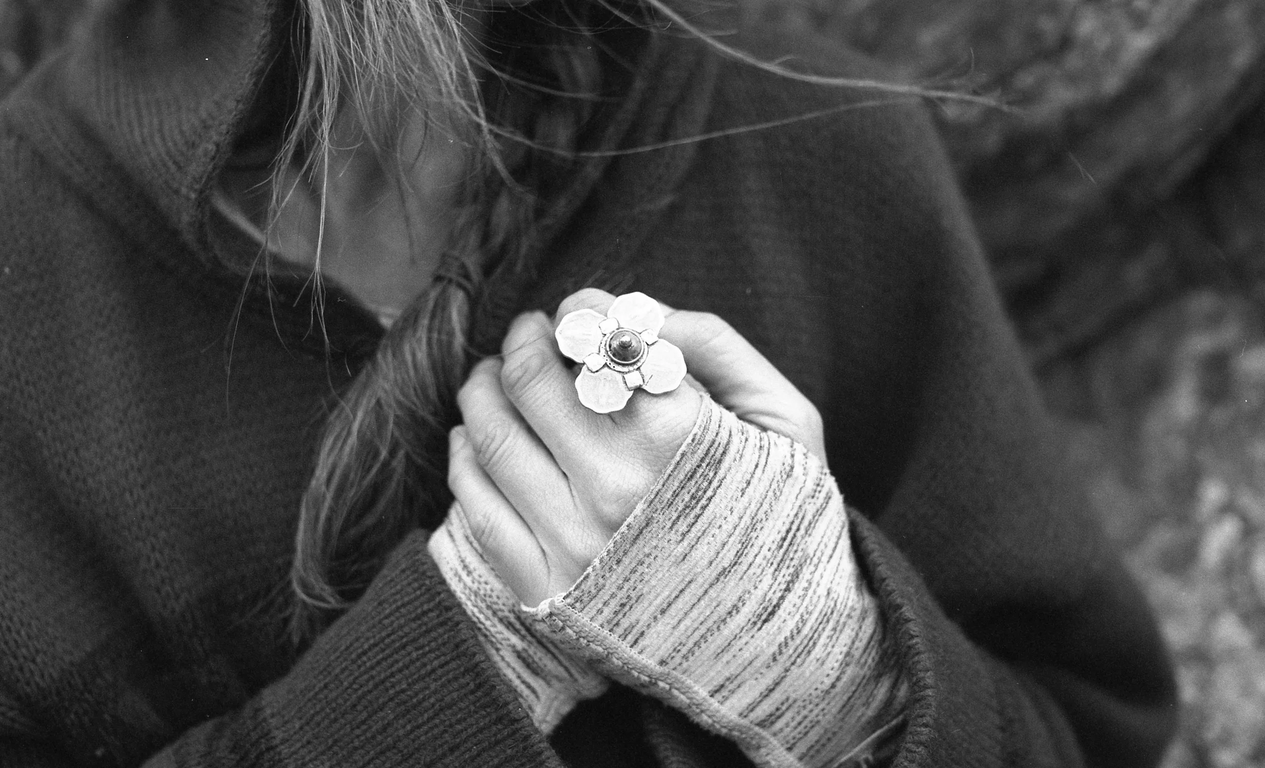Close-up of a person's hand holding a small flower-shaped object with a central gemstone, wearing a striped long-sleeve shirt and textured sweater, with long hair visible, in a black and white photograph.