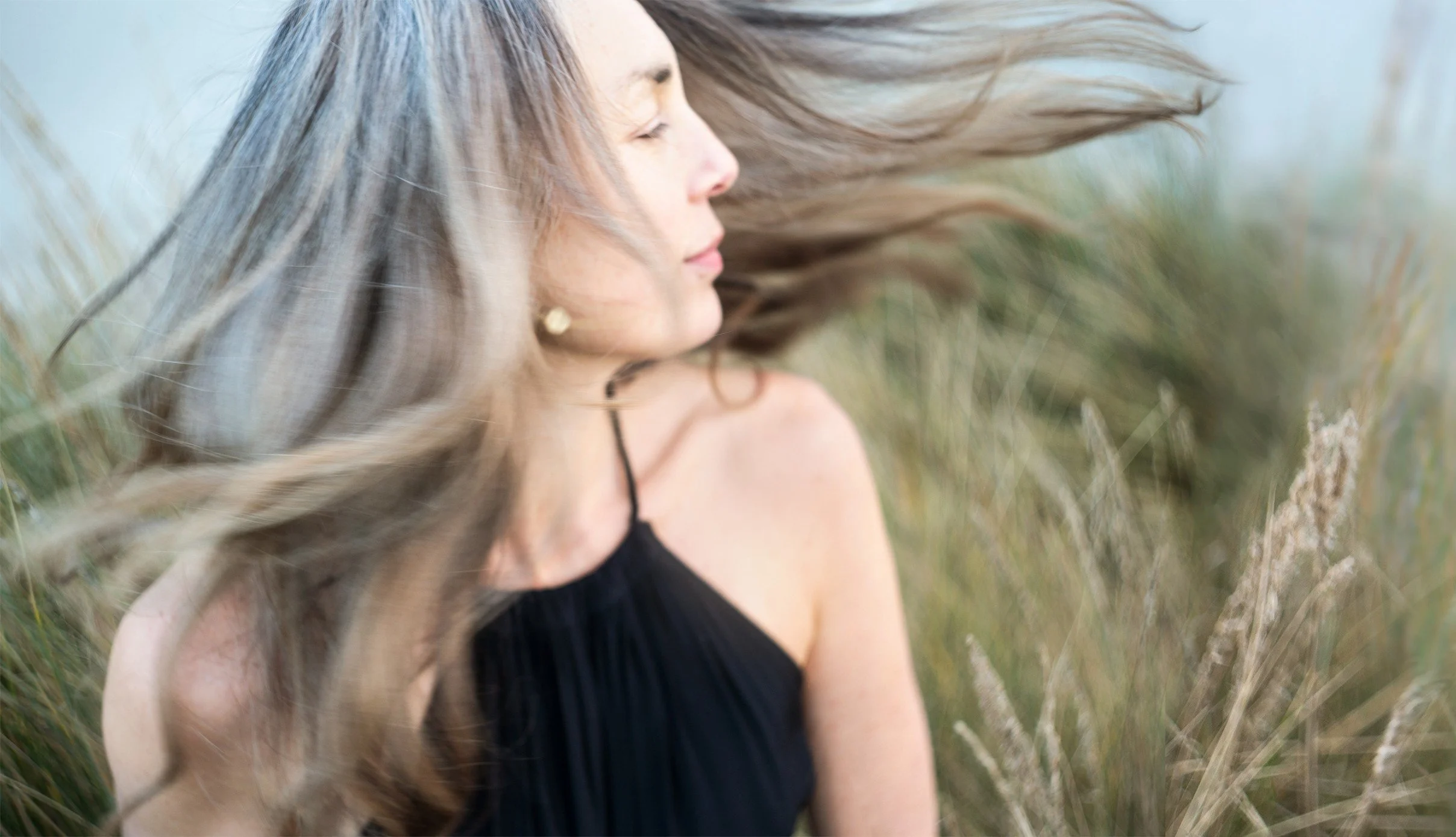 A woman with long, flowing hair appears to be outdoors with blurred grass or plants in the background, wearing a black top.