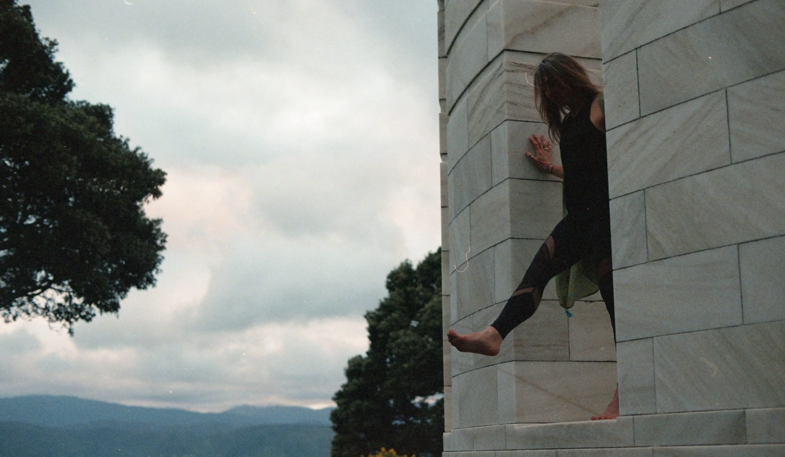 A woman in black clothing with ripped tights is sitting on a stone ledge or platform attached to a large stone building. She is leaning back with one hand on the wall and one foot hanging off the edge. Behind her, there are trees and a cloudy sky.