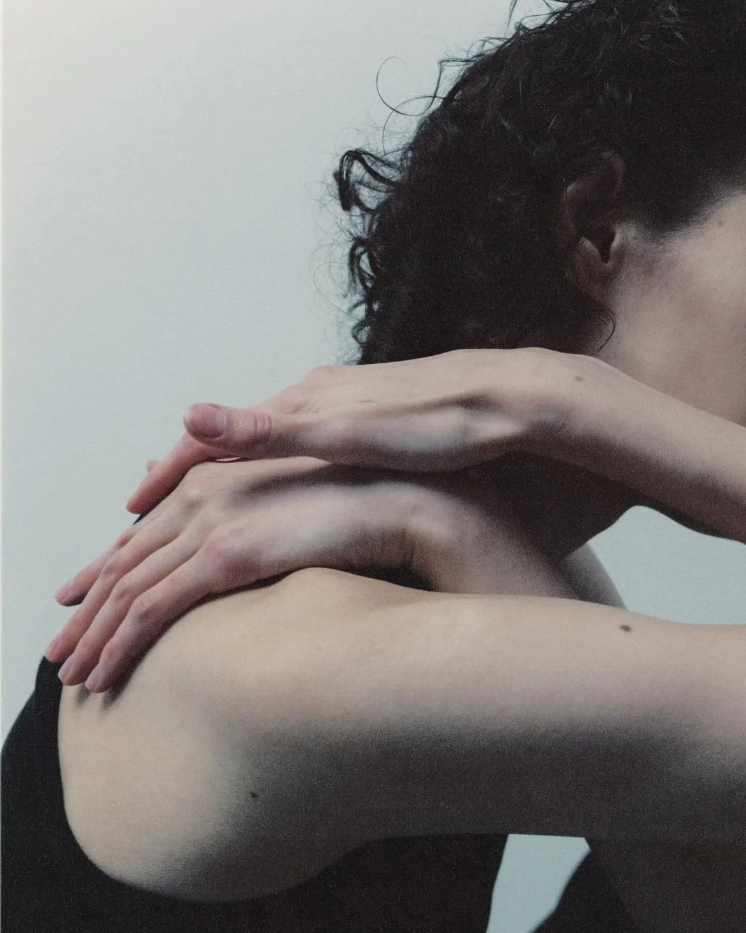 A person with curly dark hair is holding their shoulder and neck with their hands, posed against a plain light background.
