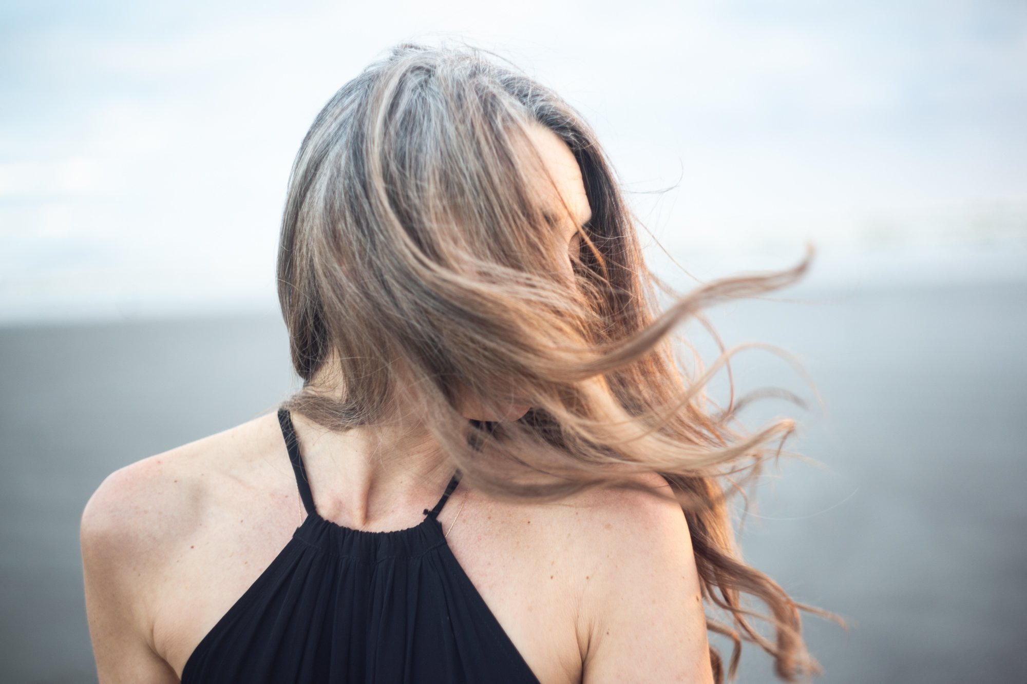 A woman with long, flowing hair wearing a black spaghetti strap top outdoors near a body of water on a cloudy day.