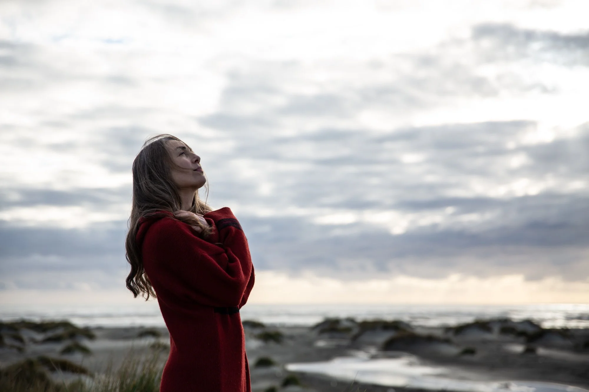 A woman with long hair in a red coat standing outdoors on a cloudy day, looking up at the sky.