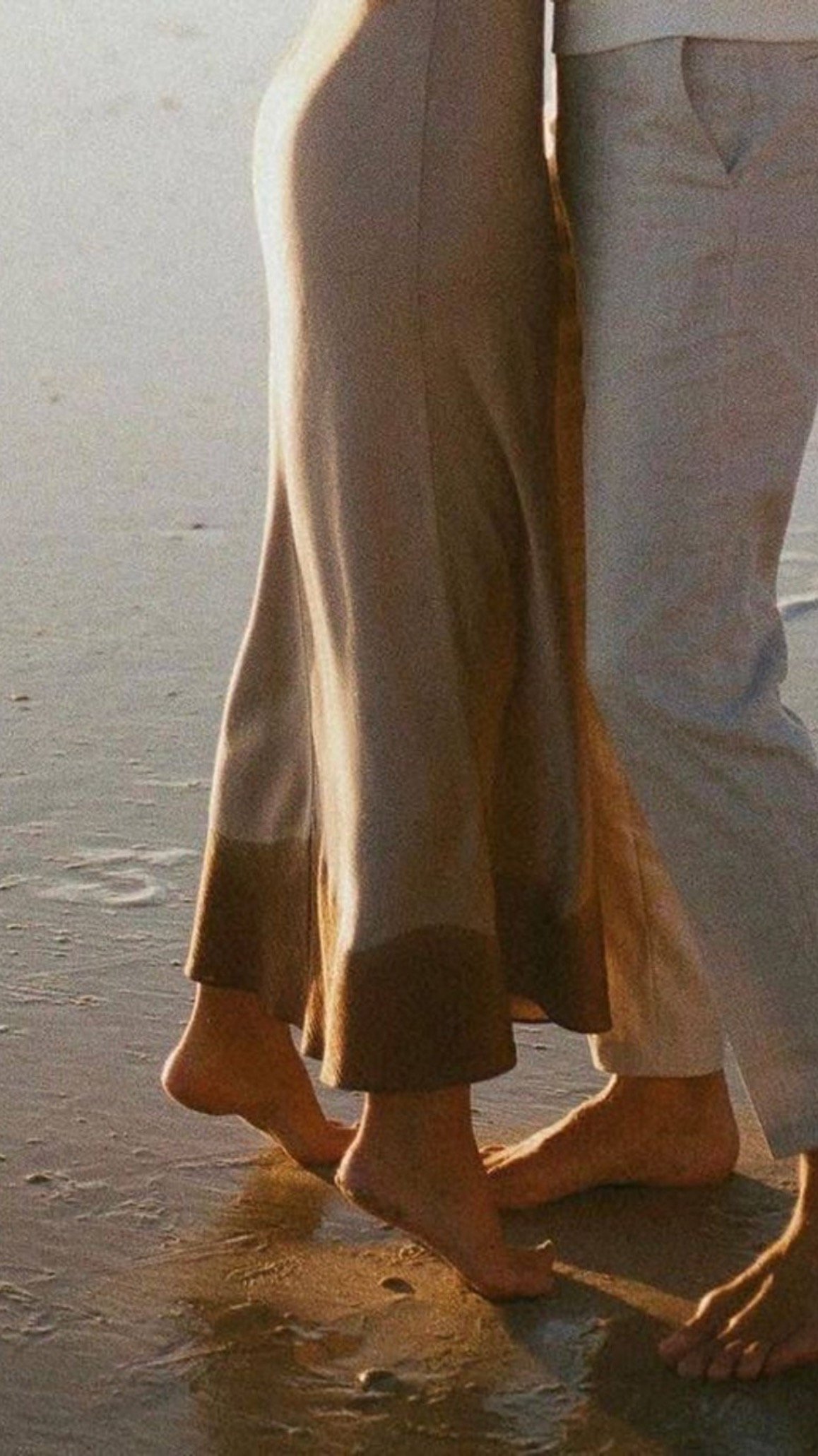 Close-up of a child barefoot on the sand with an adult, showing legs and feet at sunset or sunrise at the beach.