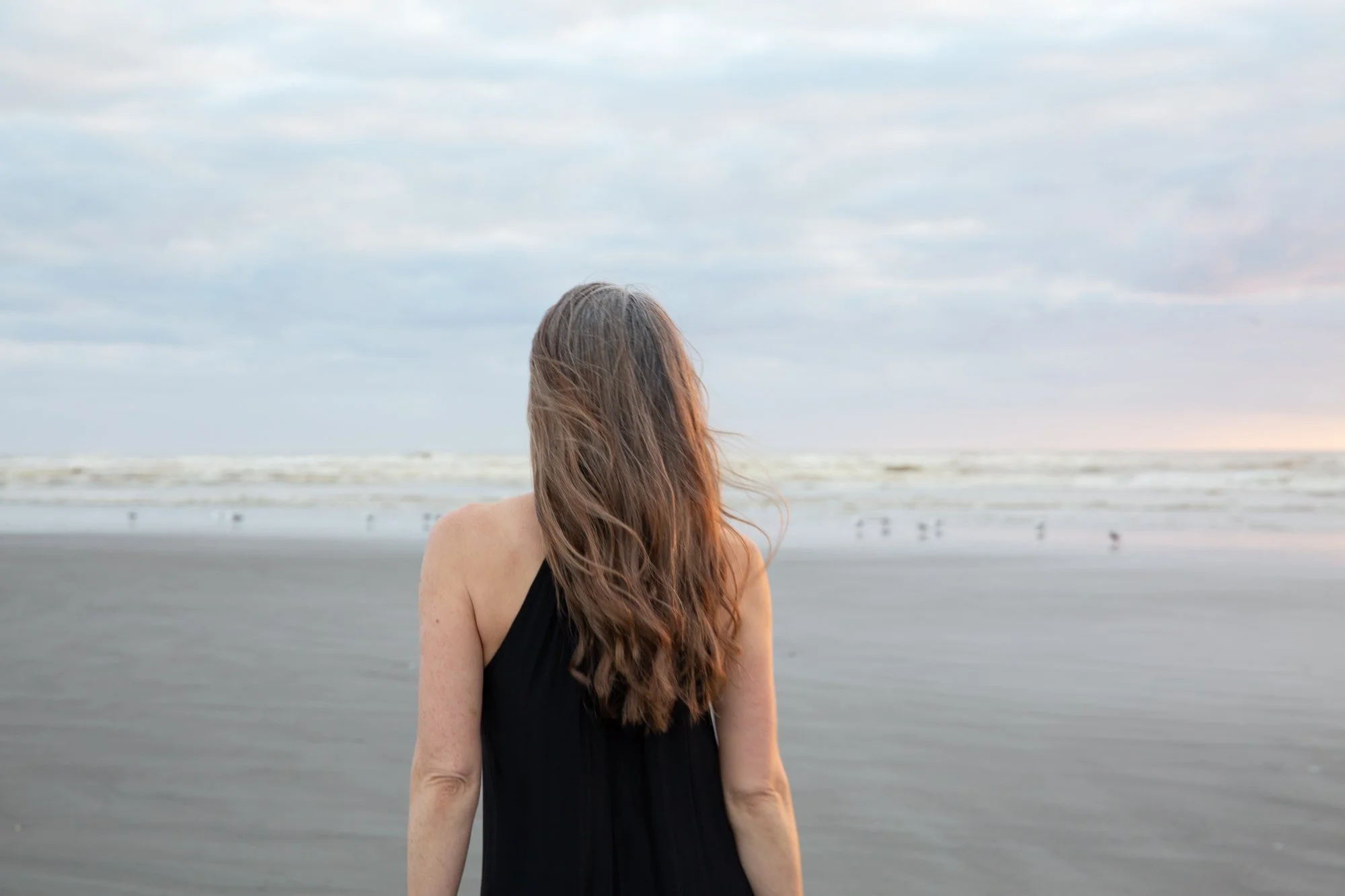 A woman with long, wavy brown hair standing on a beach, looking at the ocean during overcast weather.