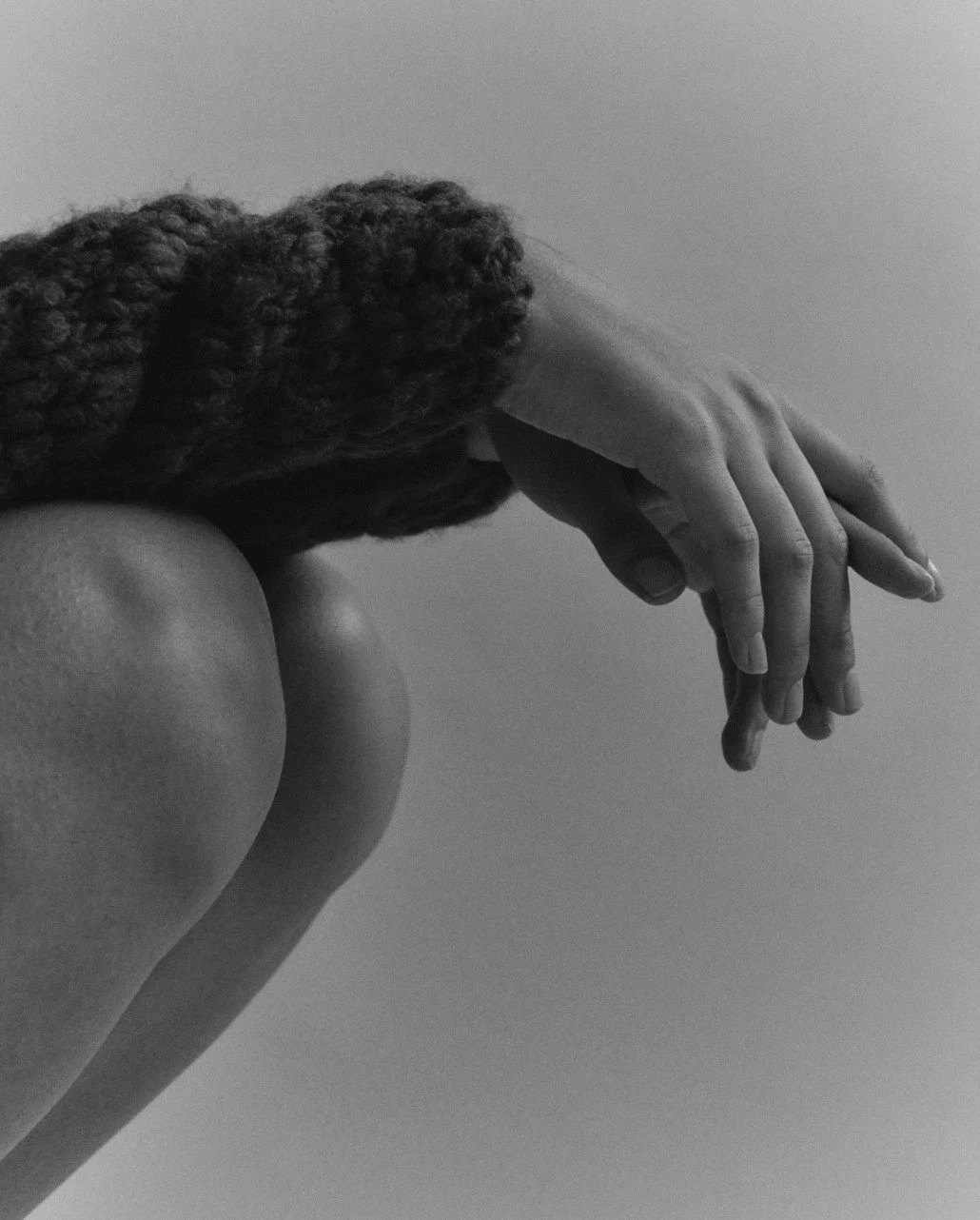A black-and-white photo of a person with curly hair resting their forehead and hand on their bent knee. The person's face is not visible.
