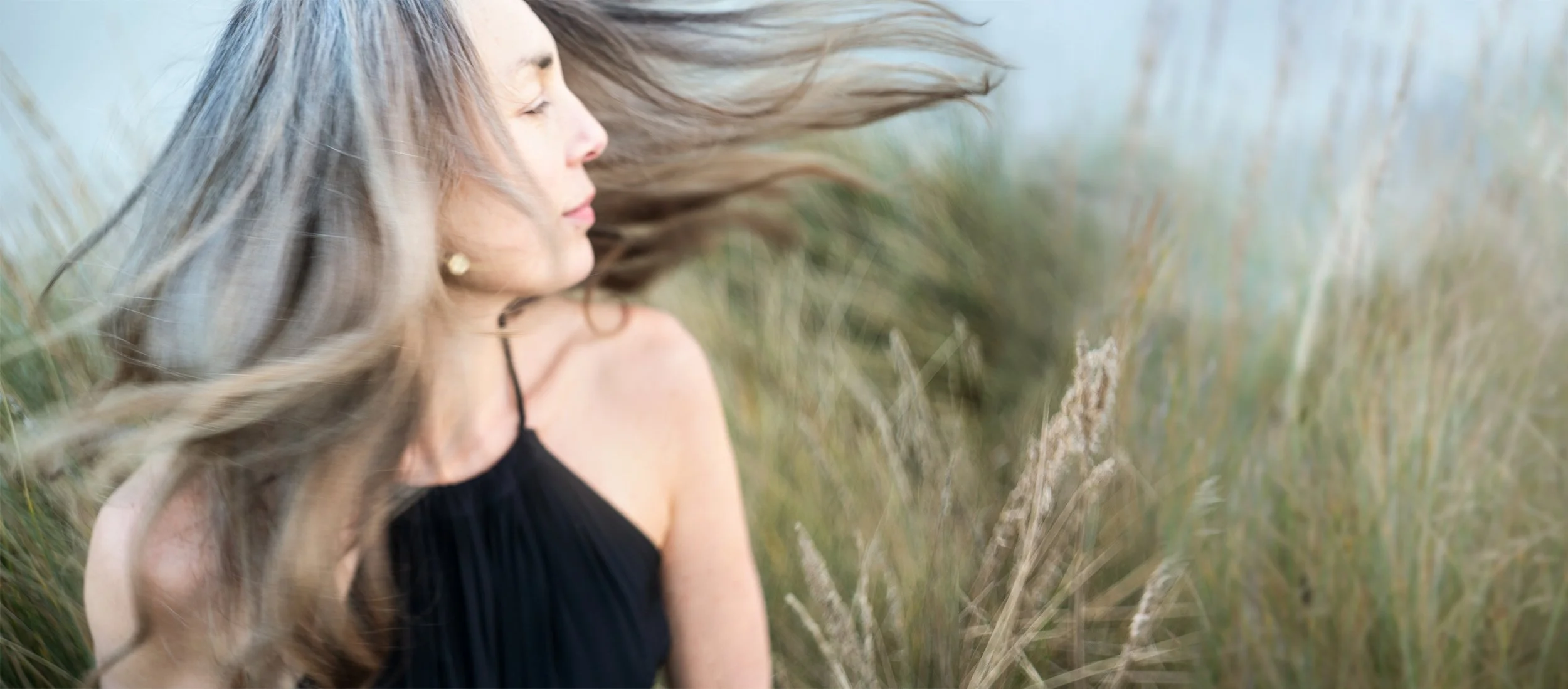 A woman with long, flowing hair is in a field of tall grass, with her eyes closed and face turned slightly to the side.