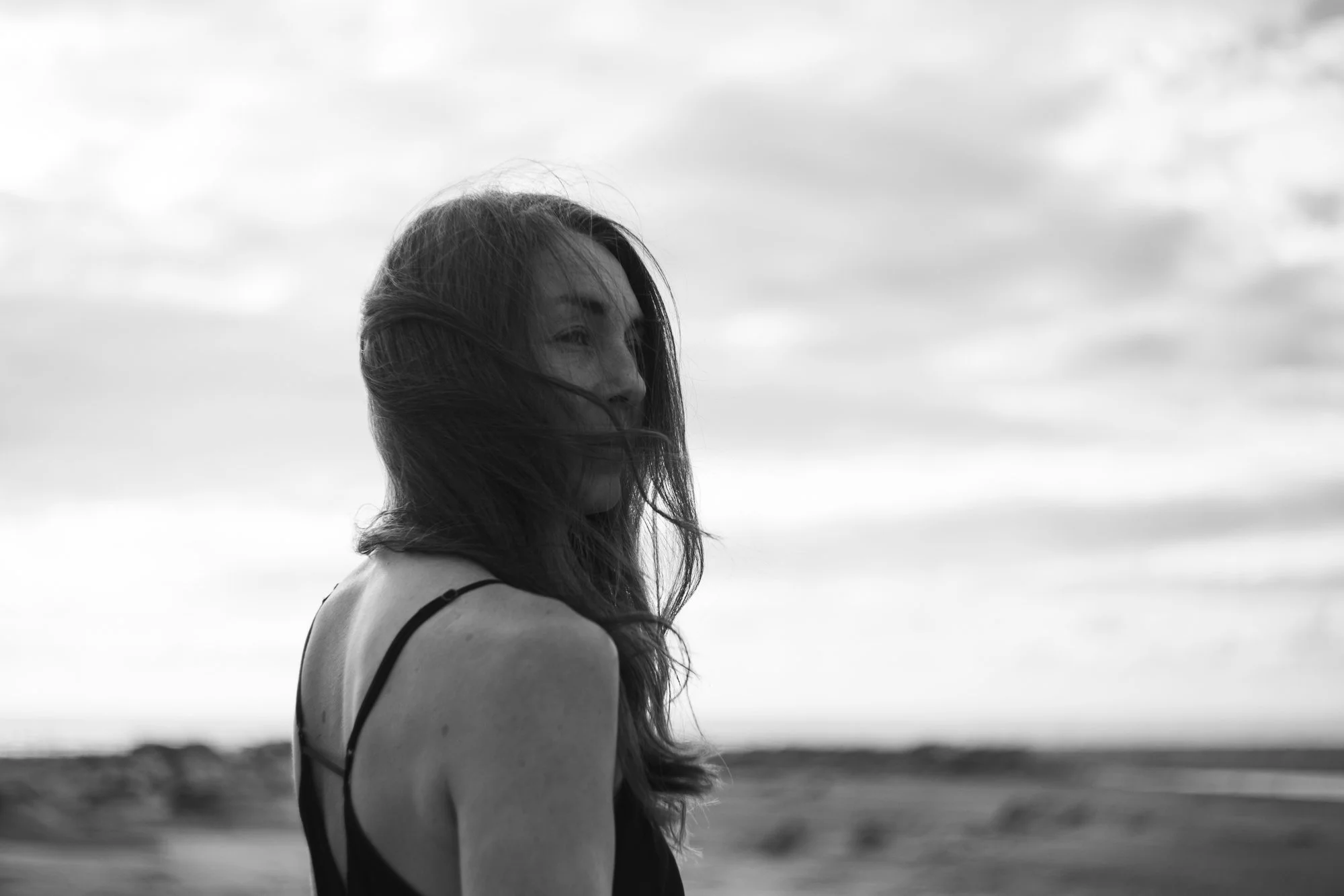 Black and white photo of a woman with long hair, standing outdoors with clouds in the sky, looking to the side.