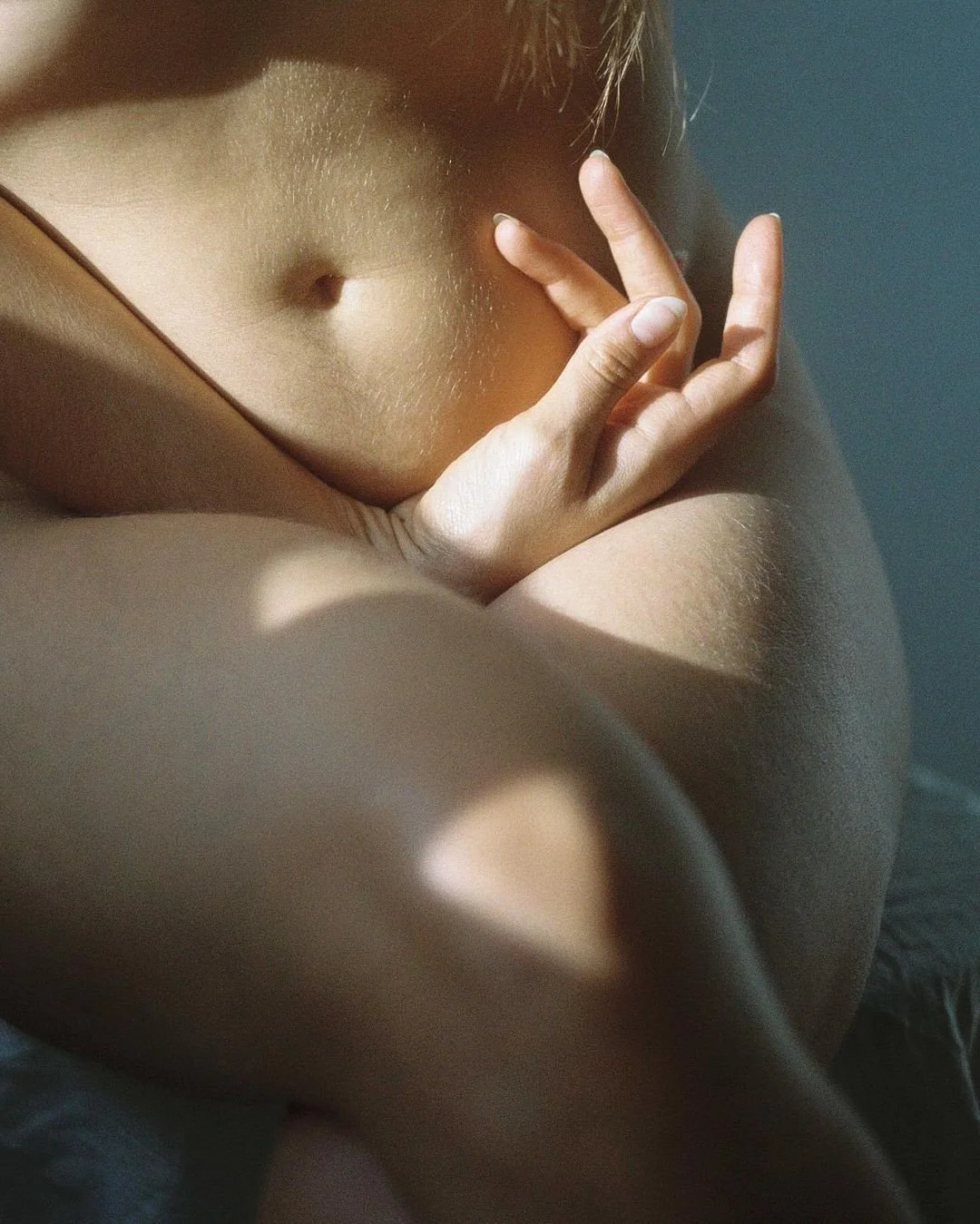 Close-up of a person's torso, showing the navel, with their hand resting on their shoulder and arm, all illuminated by soft natural light.